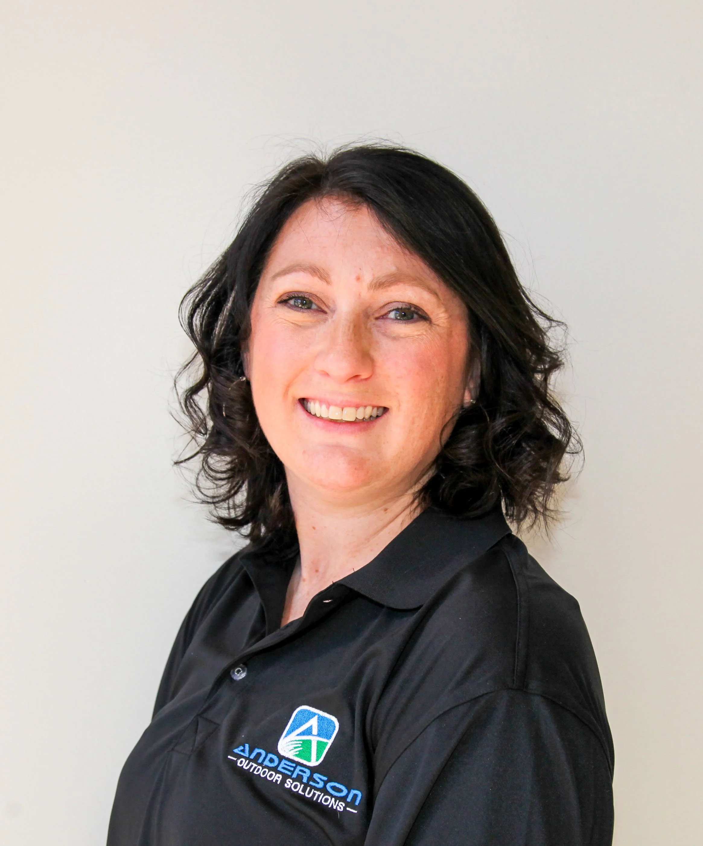 A woman with dark, curly hair smiling, wearing a black polo shirt with a logo that reads 'Anderson Outdoor Solutions'.