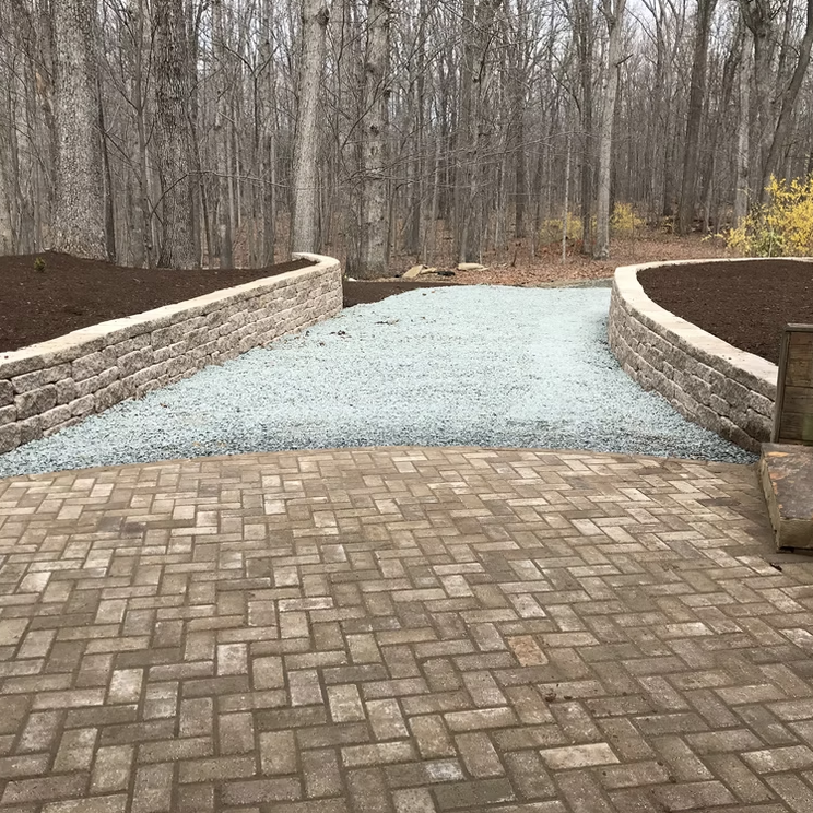 A brick patio leading to a gravel pathway flanked by stone retaining walls in a wooded backyard.