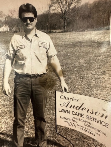 A man with dark hair and sunglasses standing next to a lawn care service sign on a grassy field with trees in the background.