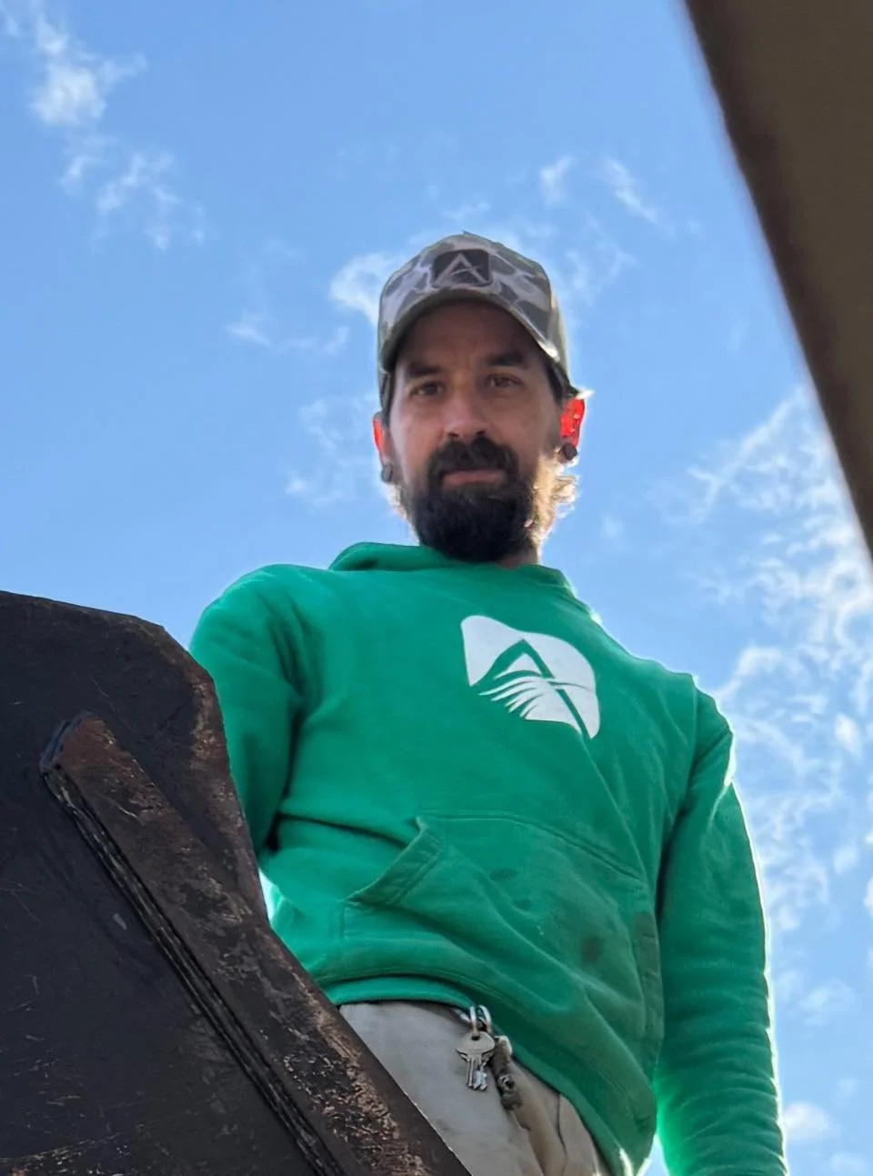 A man with a beard and mustache wearing a camouflage cap and green hoodie with a logo, looking down into the camera with a blue sky and scattered clouds in the background.