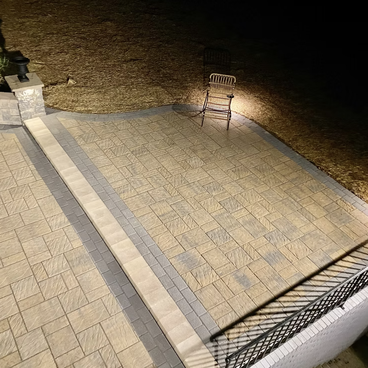 Empty outdoor patio area at night with a metal chair lit by a nearby light, surrounded by a stone and brick paved surface, and a lawn in the background.