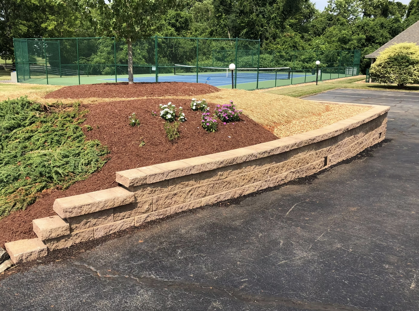A landscaped area with flowers and mulch, bordered by a stone retaining wall, adjacent to a parking lot. In the background, there is a tennis court enclosed by a green fence.