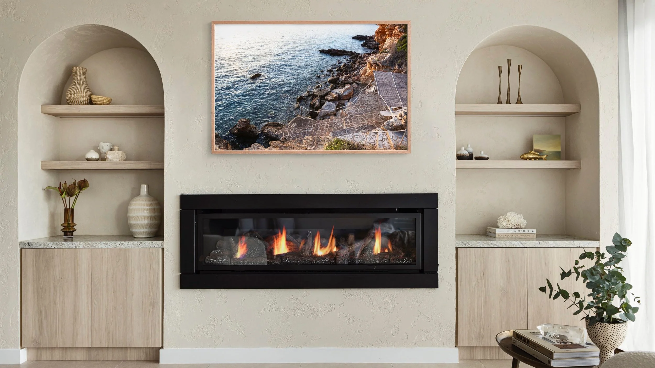 Living room with a modern fireplace, two built-in shelves with decor, a framed ocean landscape photo above the fireplace, a potted plant on a side table, and neutral-colored walls.
