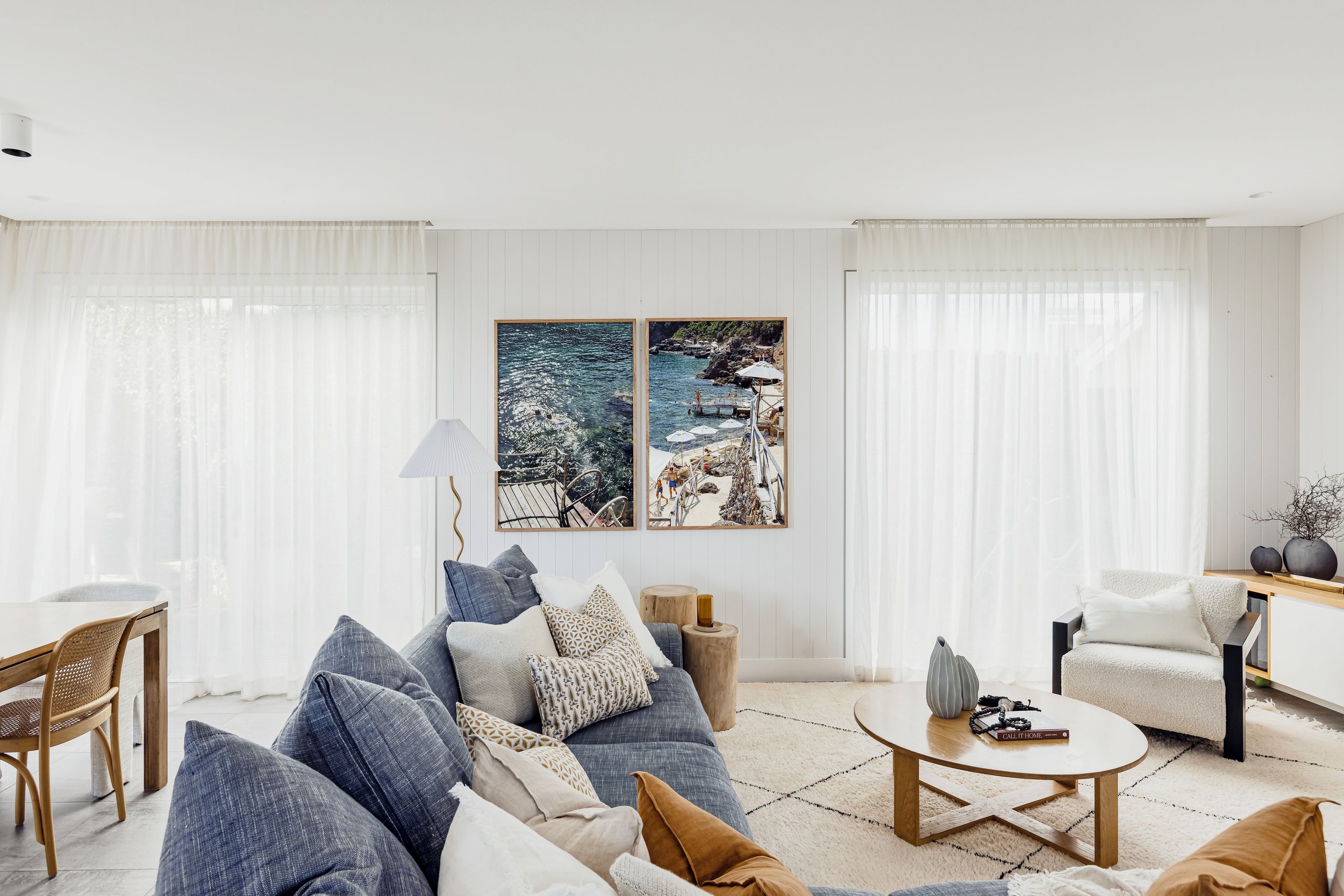 Bright living room with white walls, sheer curtains, and a variety of sofas and chairs around a round wooden coffee table. Wall art features two seaside photos.