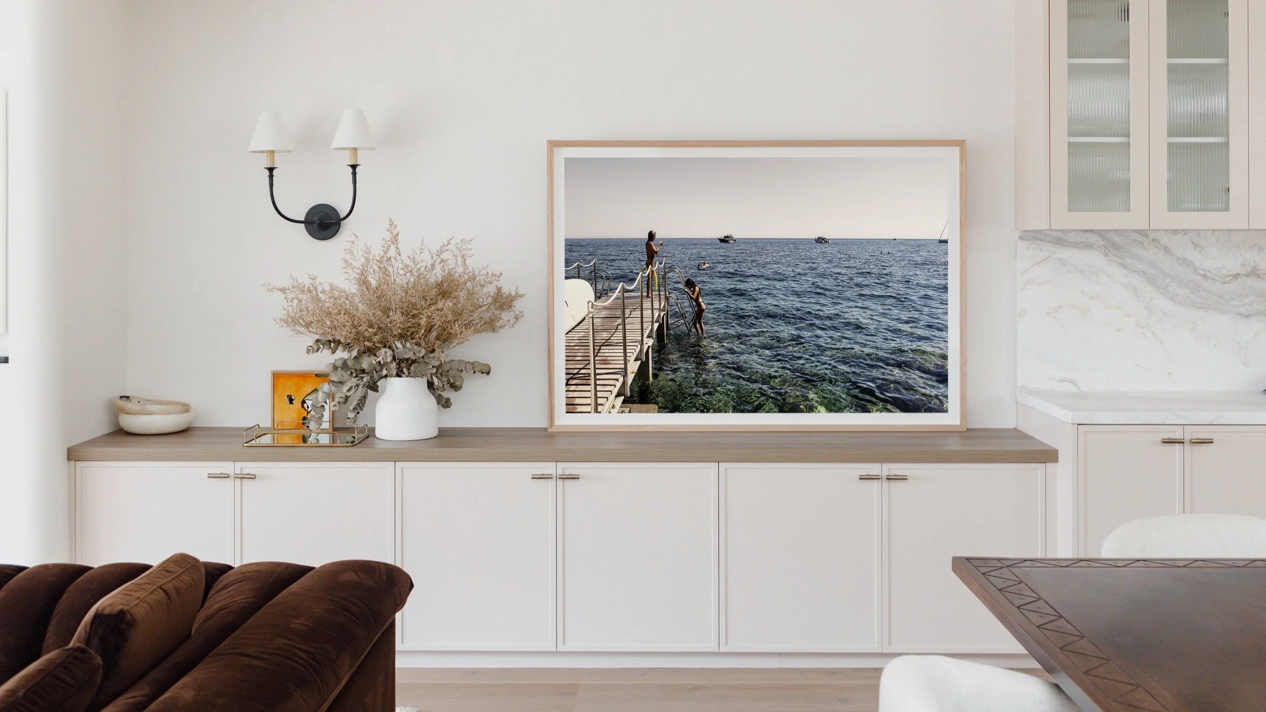 A modern living room with a white sideboard, a large framed photograph of people on a pier by the ocean, dried flowers in a white vase, and a wall-mounted lamp with two shades.