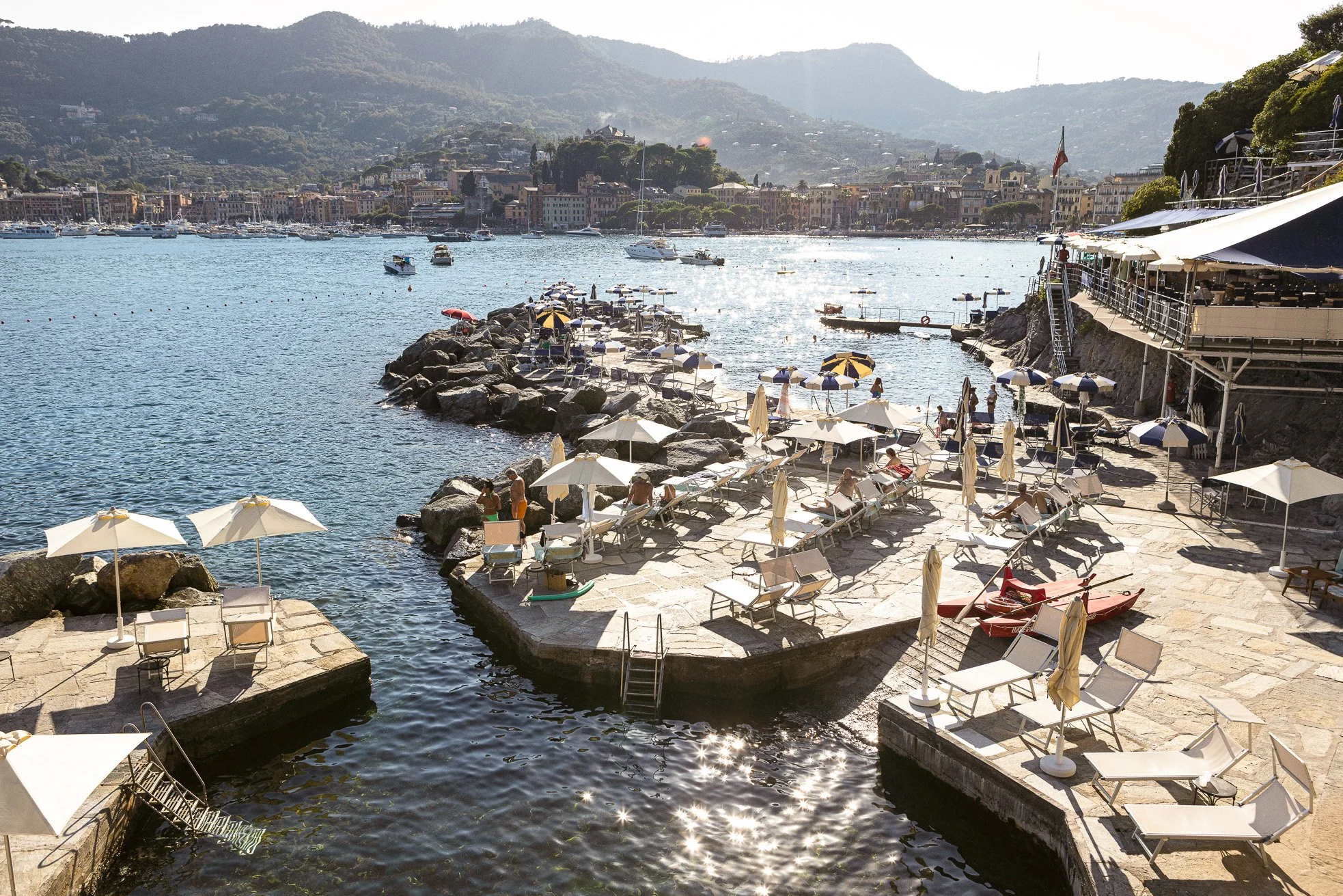 A seaside area with lounge chairs and umbrellas along the rocky shoreline, people relaxing and sunbathing, boats anchored in the water, and a cityscape with buildings and hills in the background.