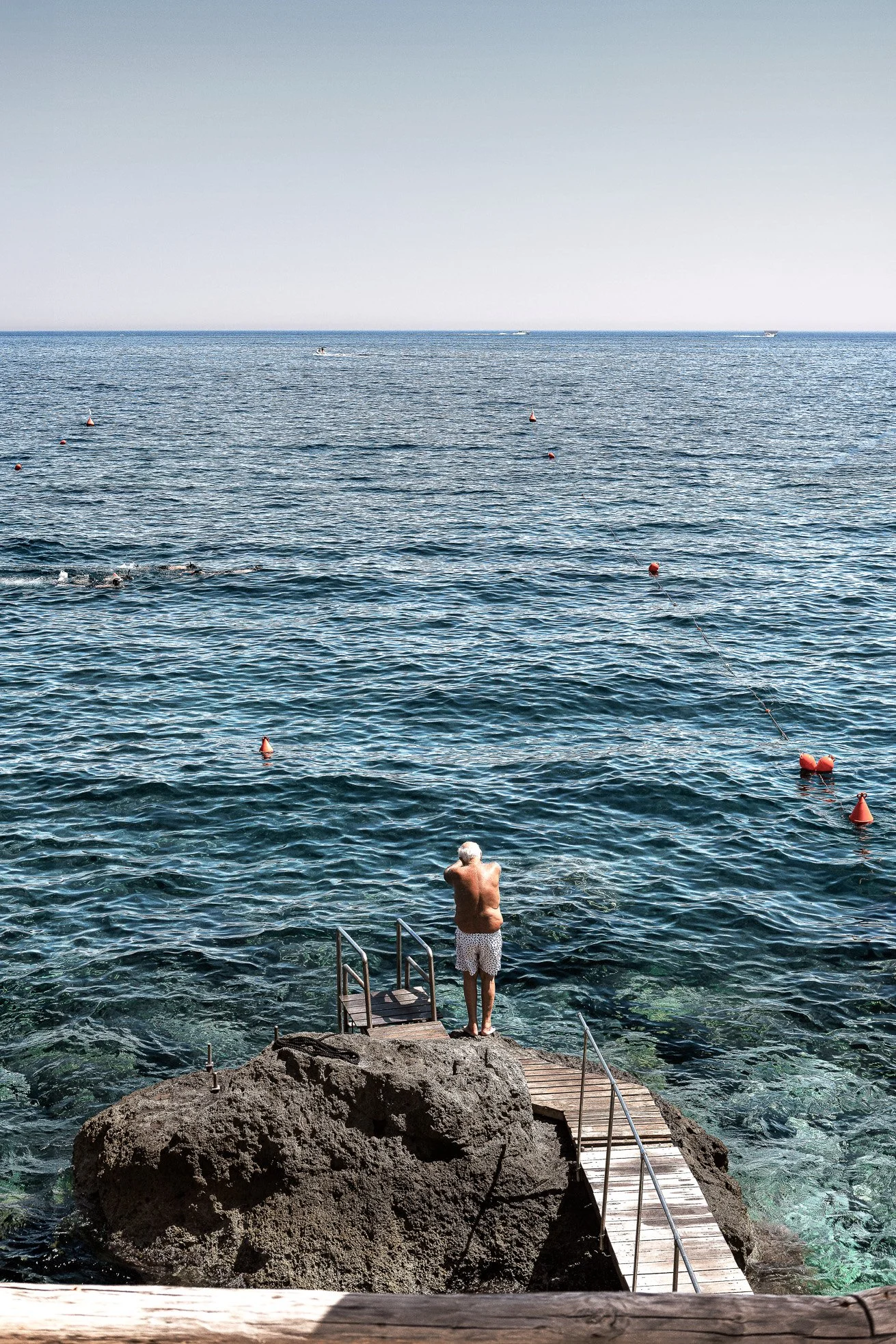 An elderly man standing on a rocky platform at the edge of the water, looking out over the ocean. The platform is connected to a wooden walkway with railings. The ocean has small waves and several buoys floating, with boats in the distance under a cl