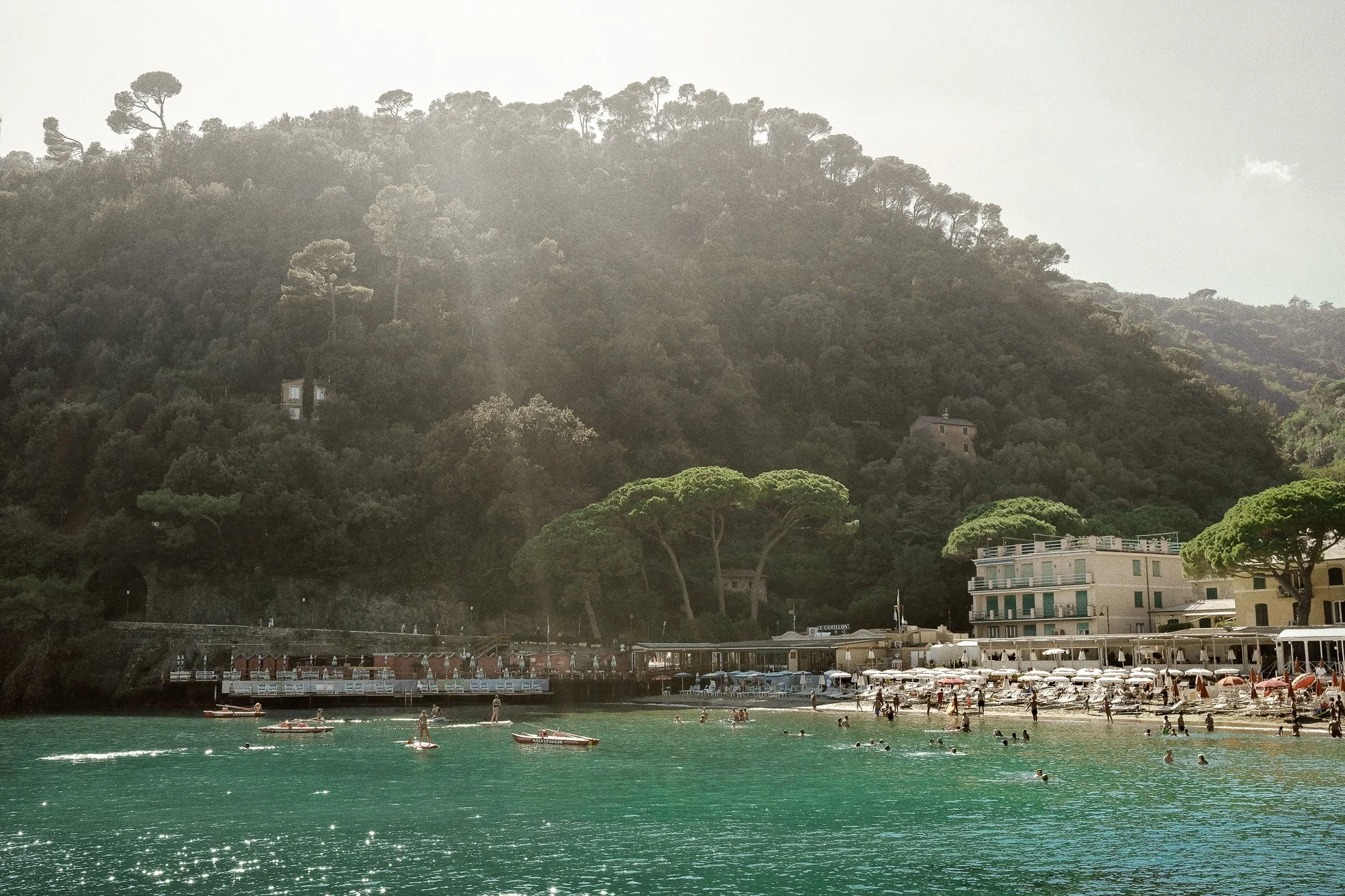 Beach with umbrellas and people swimming, backed by a hillside with trees and buildings, in sunlight.