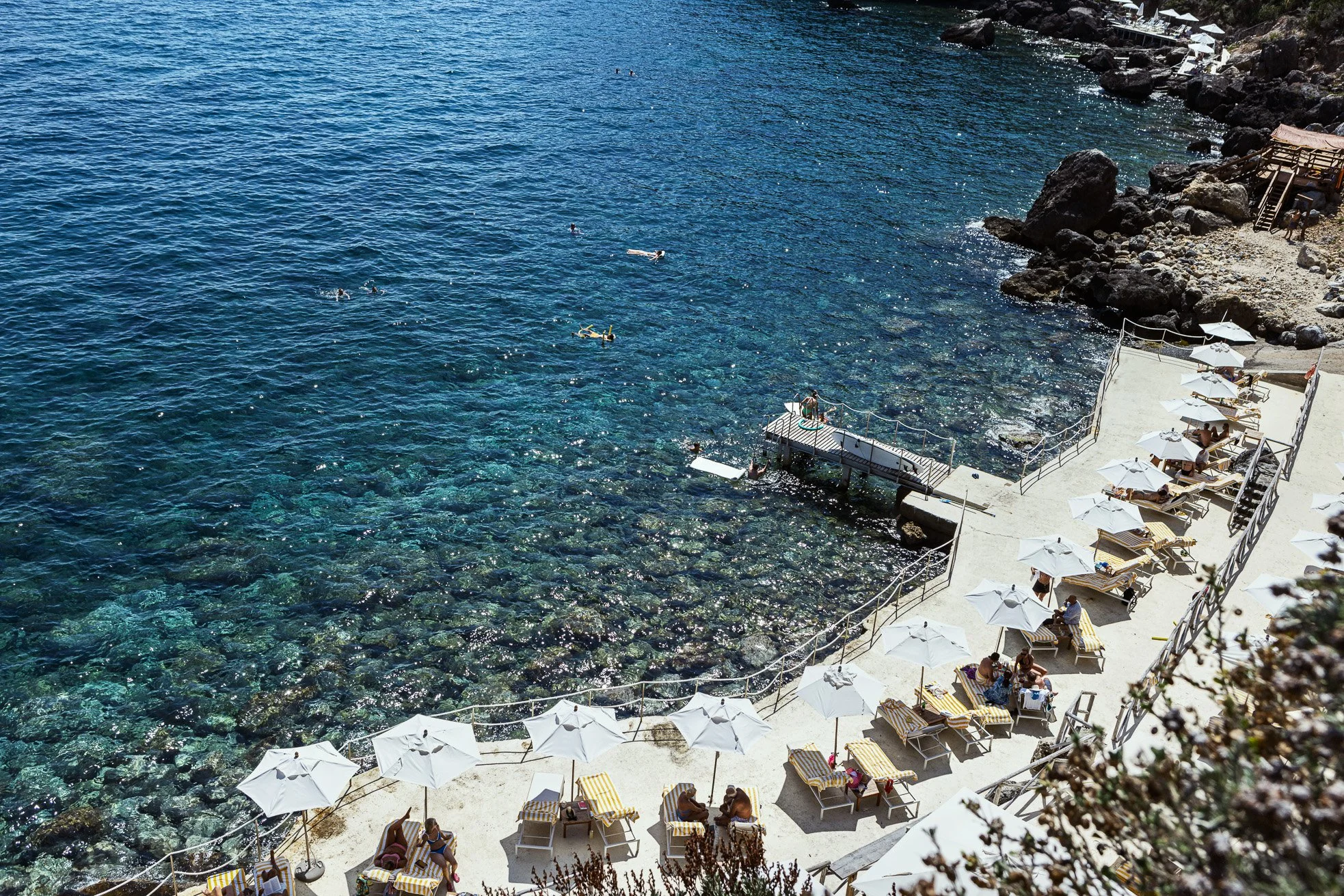 A sunny beachside scene with a concrete deck and white umbrellas. People are sunbathing on lounge chairs and swimming in the clear blue water near the rocky shoreline.