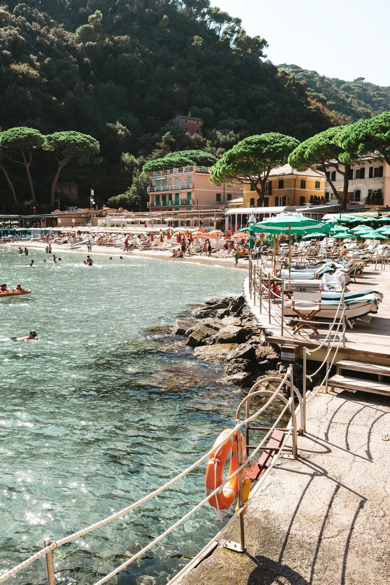 Beach scene with people swimming and lounging under umbrellas, rocky shoreline, colorful buildings, and green mountains in the background.