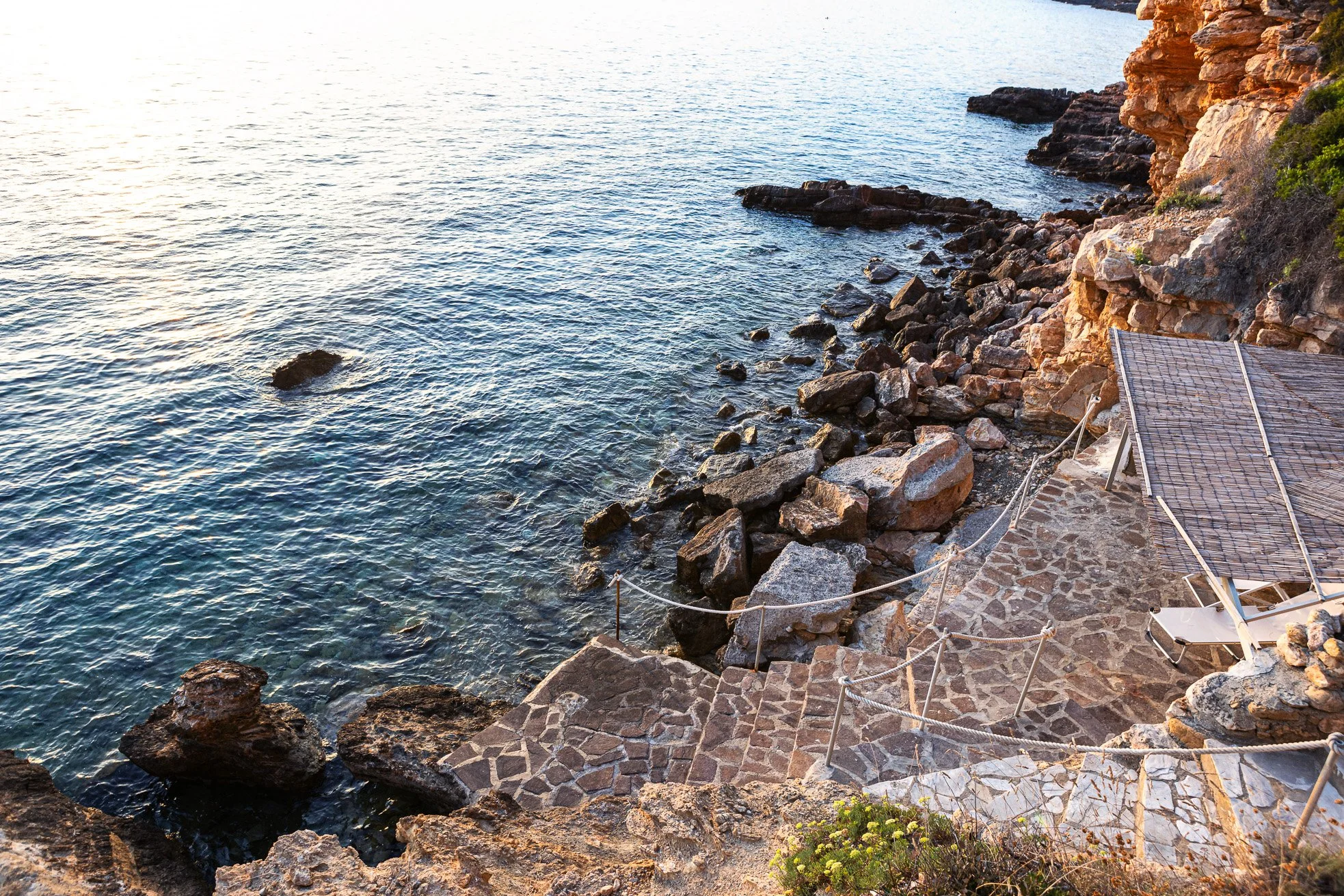 A rocky seaside with a stone pathway and a sun lounge chair next to the water, a cliff with lush greenery, and the ocean reflecting sunset light.