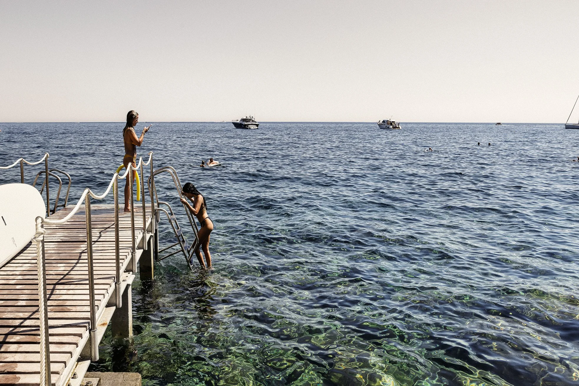 People swimming and standing on a dock by the ocean with boats in the background and clear water in the foreground.