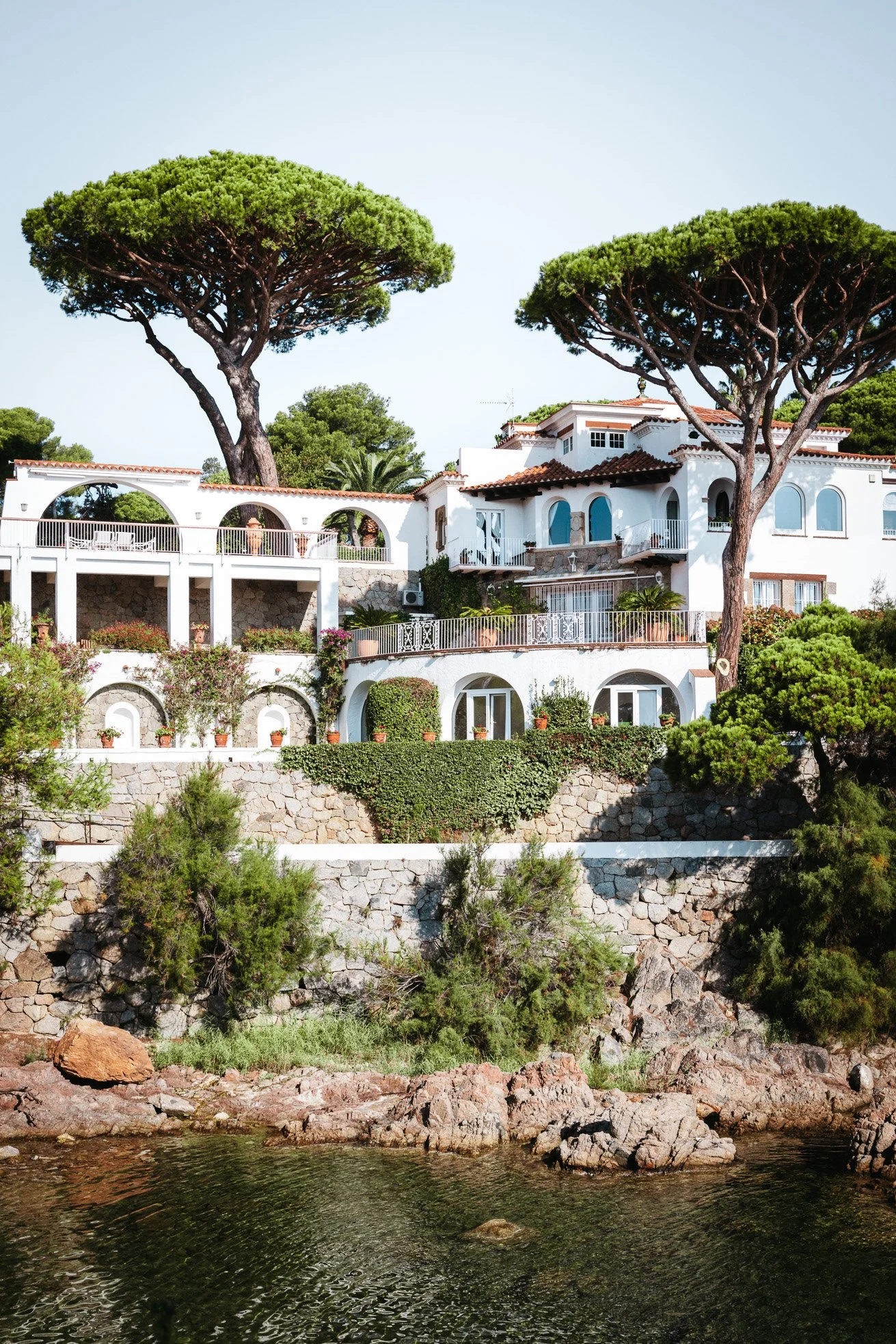 White Mediterranean-style house with terracotta roof tiles, surrounded by lush green trees and bushes, overlooking a rocky shoreline with water.