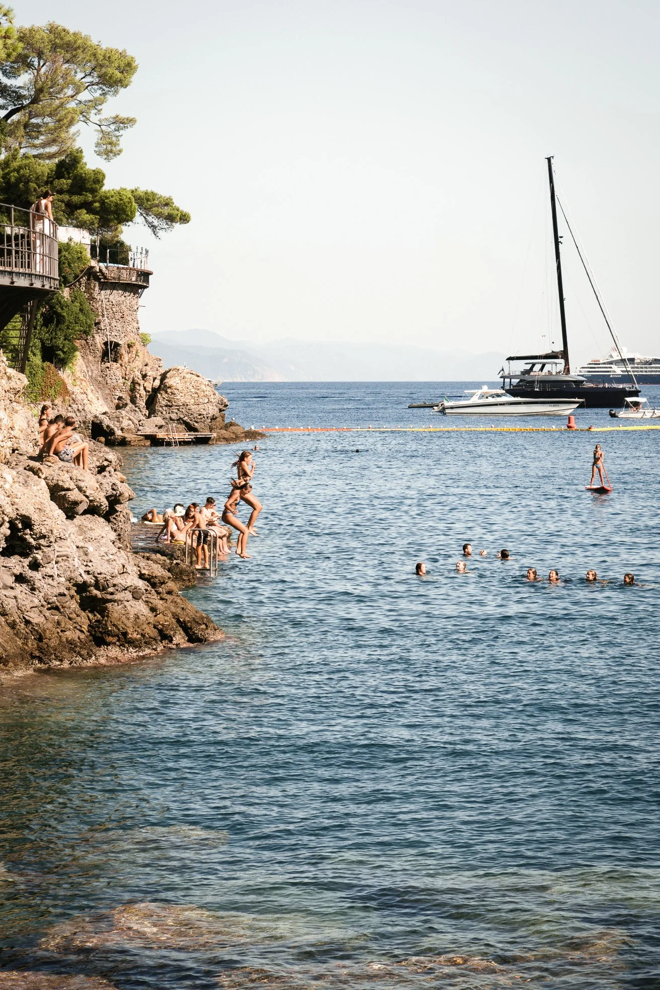 People swimming and relaxing by a rocky seaside with boats anchored nearby, and a person paddleboarding on the water.