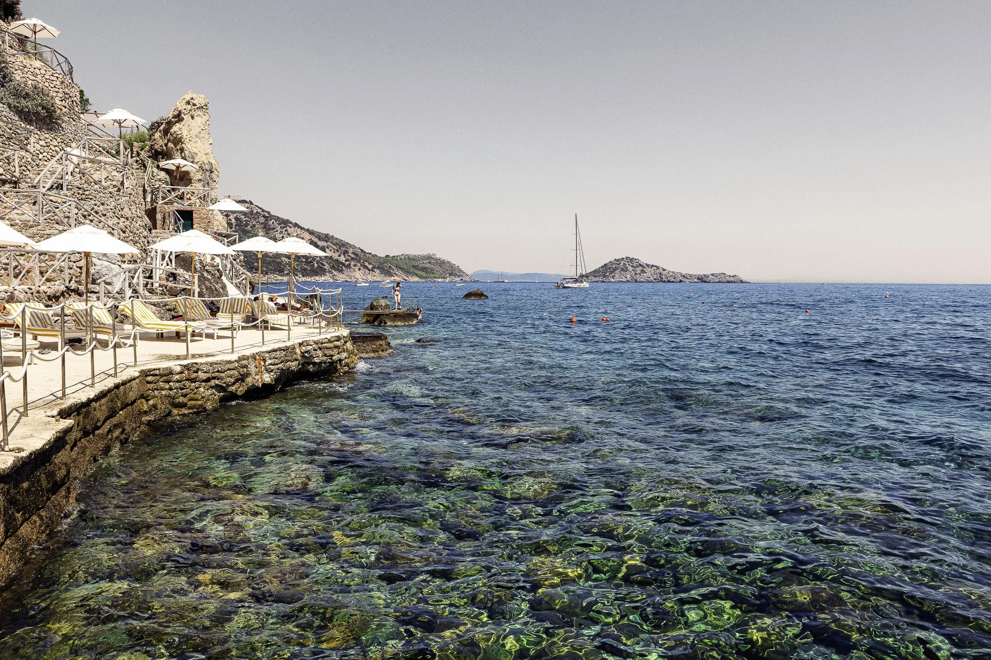 Seaside view with rocky coastline, lounge chairs with yellow and white striped cushions, white umbrellas, and a calm ocean with a sailboat in the distance.