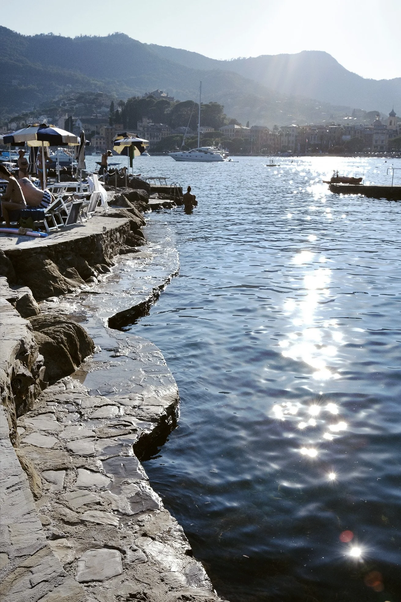 People relaxing on lounge chairs under umbrellas by the rocky waterfront, with boats on the water and a hillside town in the background on a sunny day.