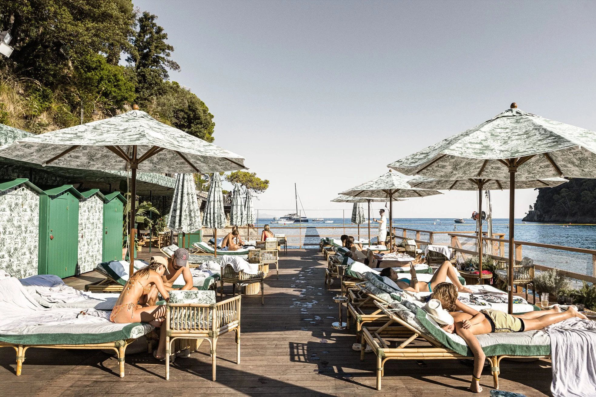 People relaxing on lounge chairs and under umbrellas on a wooden deck by the ocean, with boats in the background and a hillside on the left.