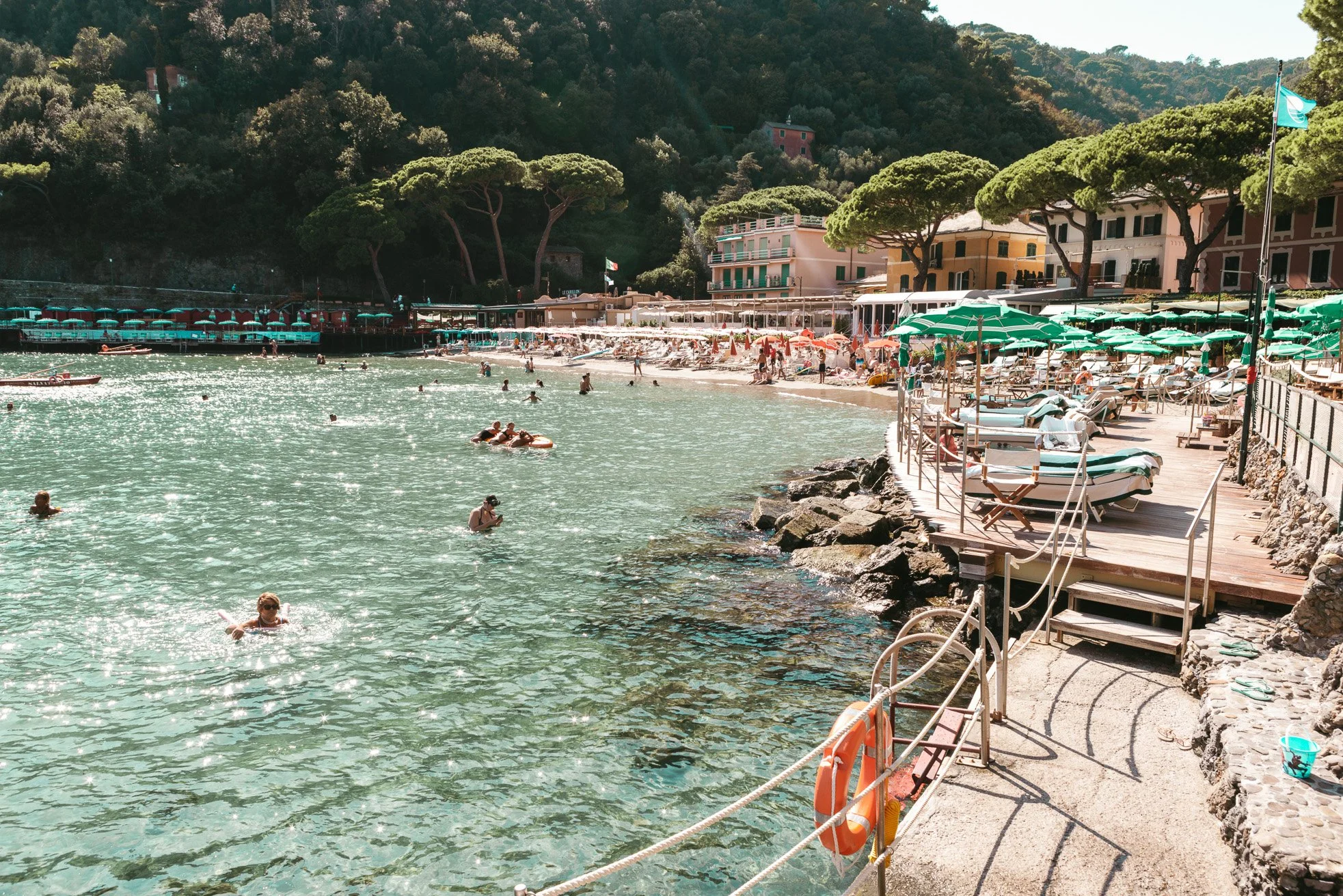 Coastal beach scene with people swimming in clear water, surrounded by colorful umbrellas and sun loungers along a rocky shoreline; buildings and lush green trees in the background.