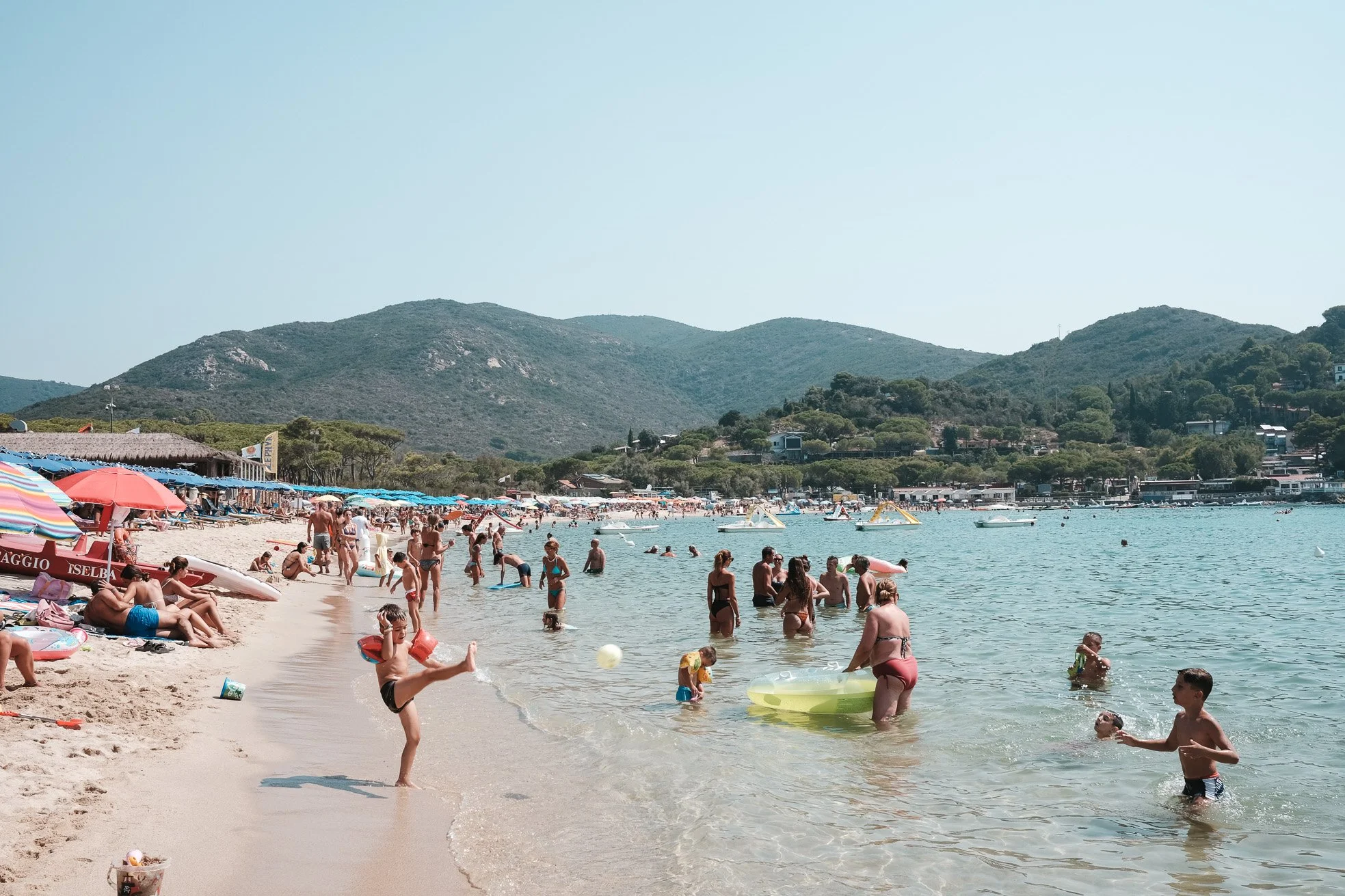 Beach scene with people swimming, playing, and relaxing on the shore, under umbrellas, with hills in the background.