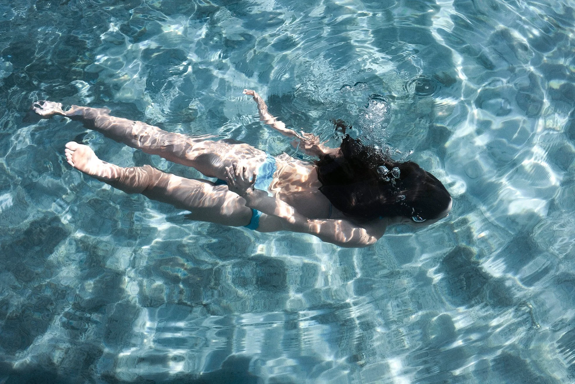 A person with long dark hair floating face down in a swimming pool, wearing a black swimsuit and light-colored swim trunks, with ripples and sunlight reflections on the water surface.