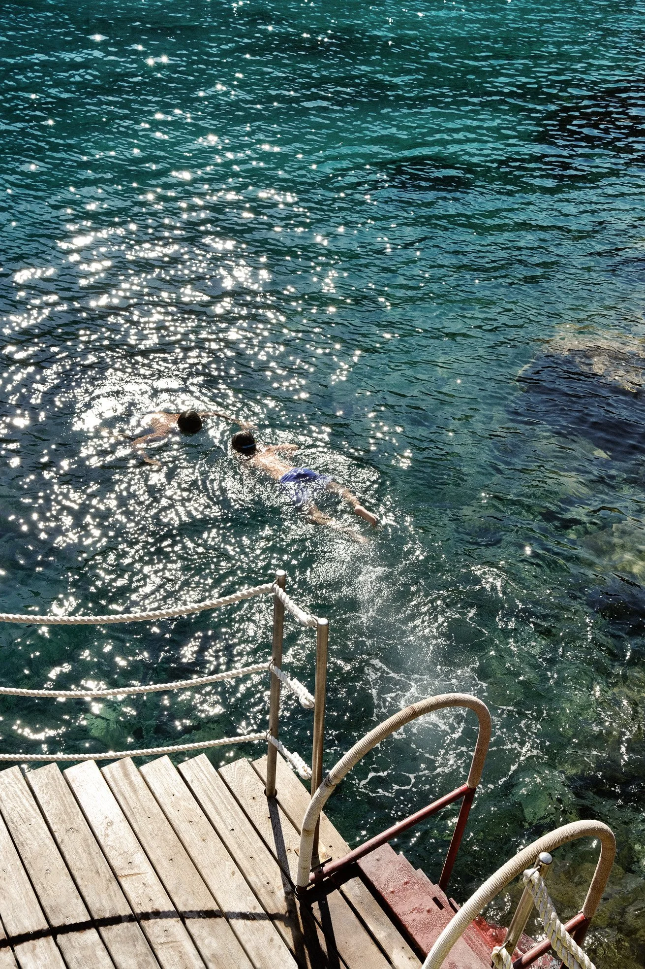 Two people swimming in clear water near a wooden dock with a railing.
