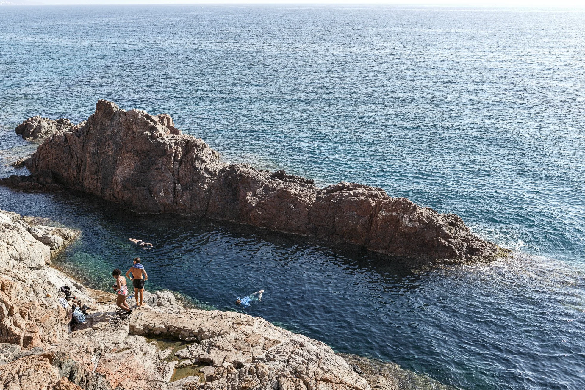 People swimming and relaxing near rocky coast and tide pool with a large rock formation by the ocean.