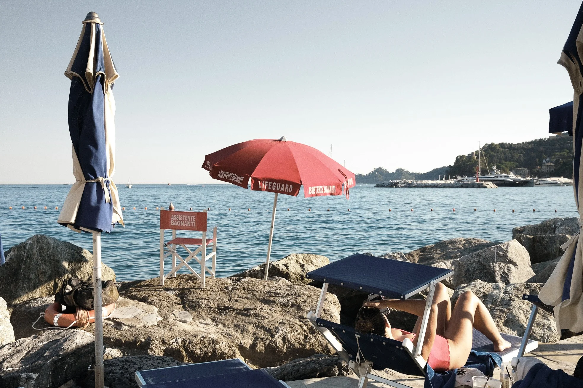 Beach scene with a woman sunbathing on a lounger near the rocky shore, surrounded by umbrellas, with a red umbrella marked 'ASSISTENTE BAGNANTI' and a red chair labeled 'ASSISTENTE BAGNANTI', boat in the water, and a distant coastline with greenery a