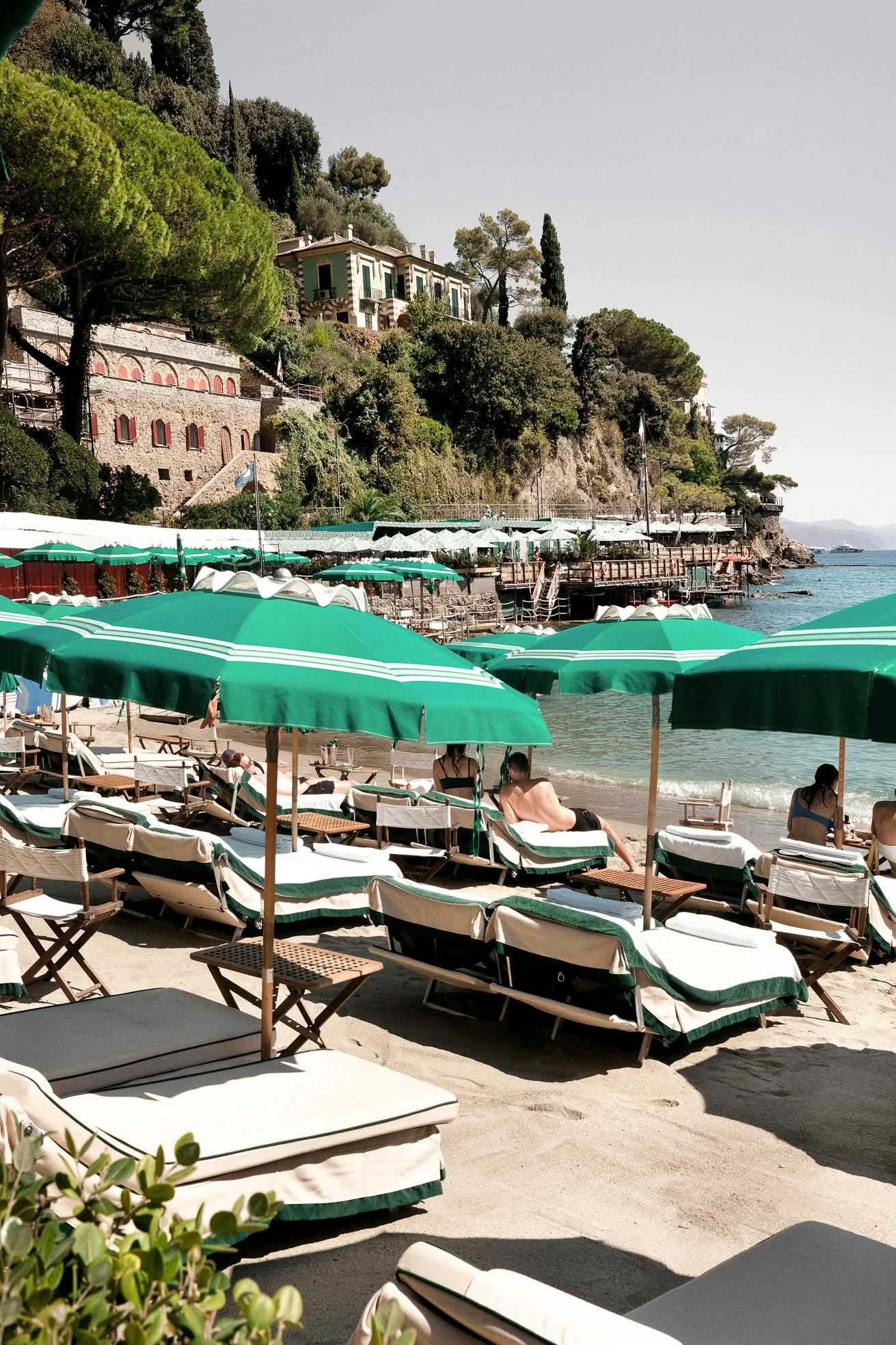 Beach with lounge chairs and green umbrellas near the water, against a hillside with houses and trees.