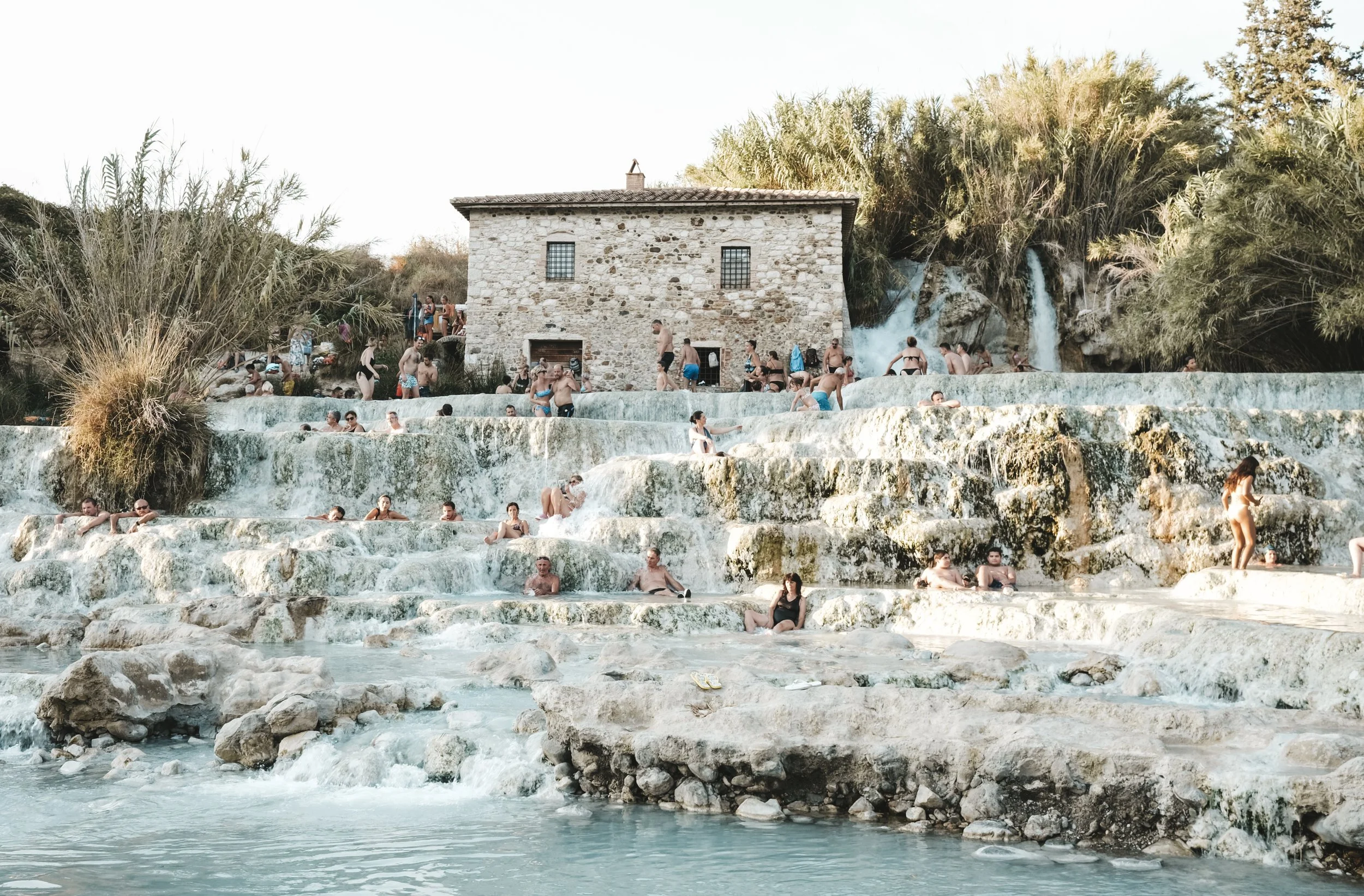 People relaxing and swimming in natural hot springs with a stone building and lush vegetation in the background.