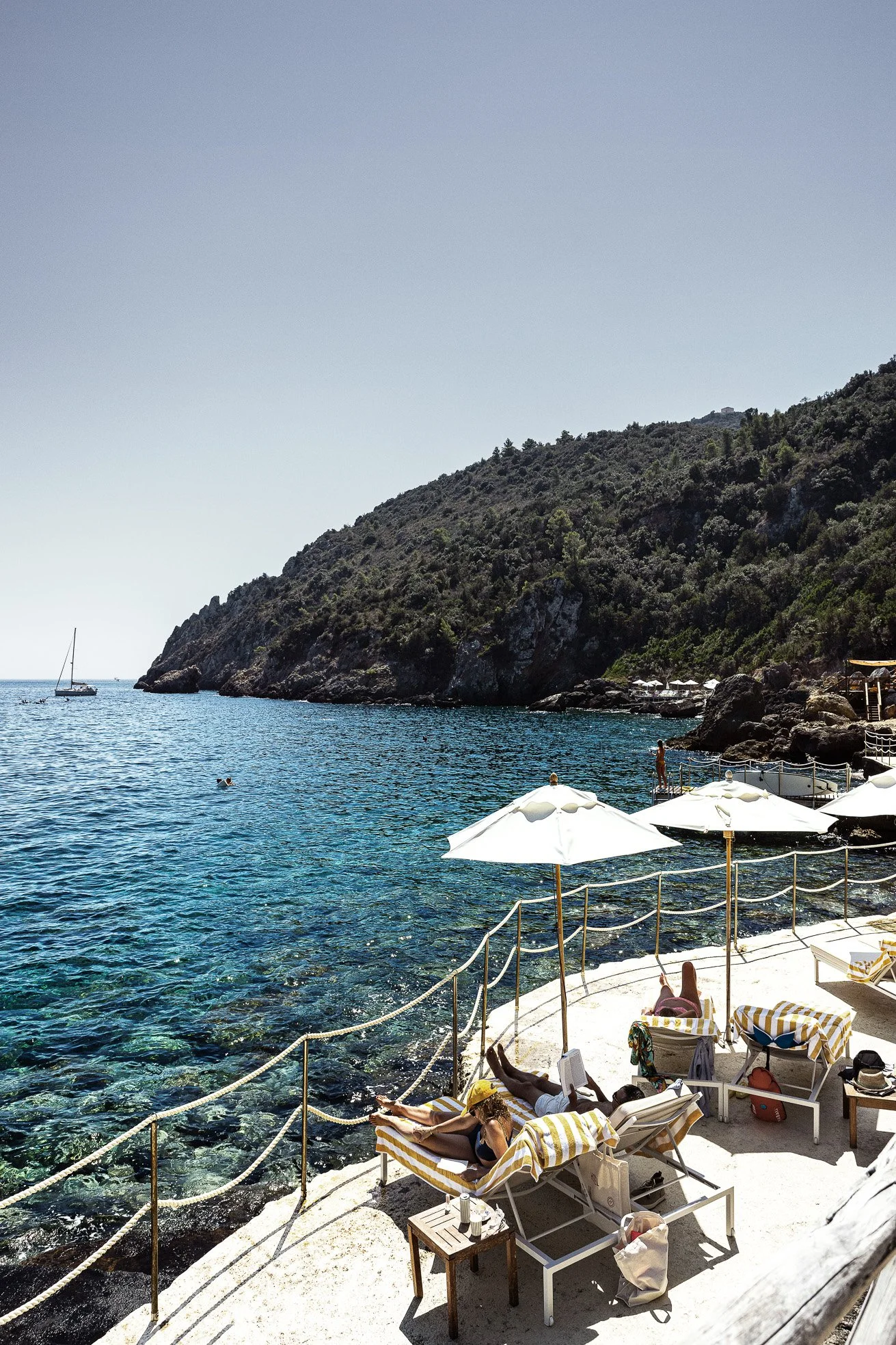 People relaxing on lounge chairs with yellow and white striped towels on a rocky seaside patio with white umbrellas, overlooking blue water and a sailboat near a green hillside.