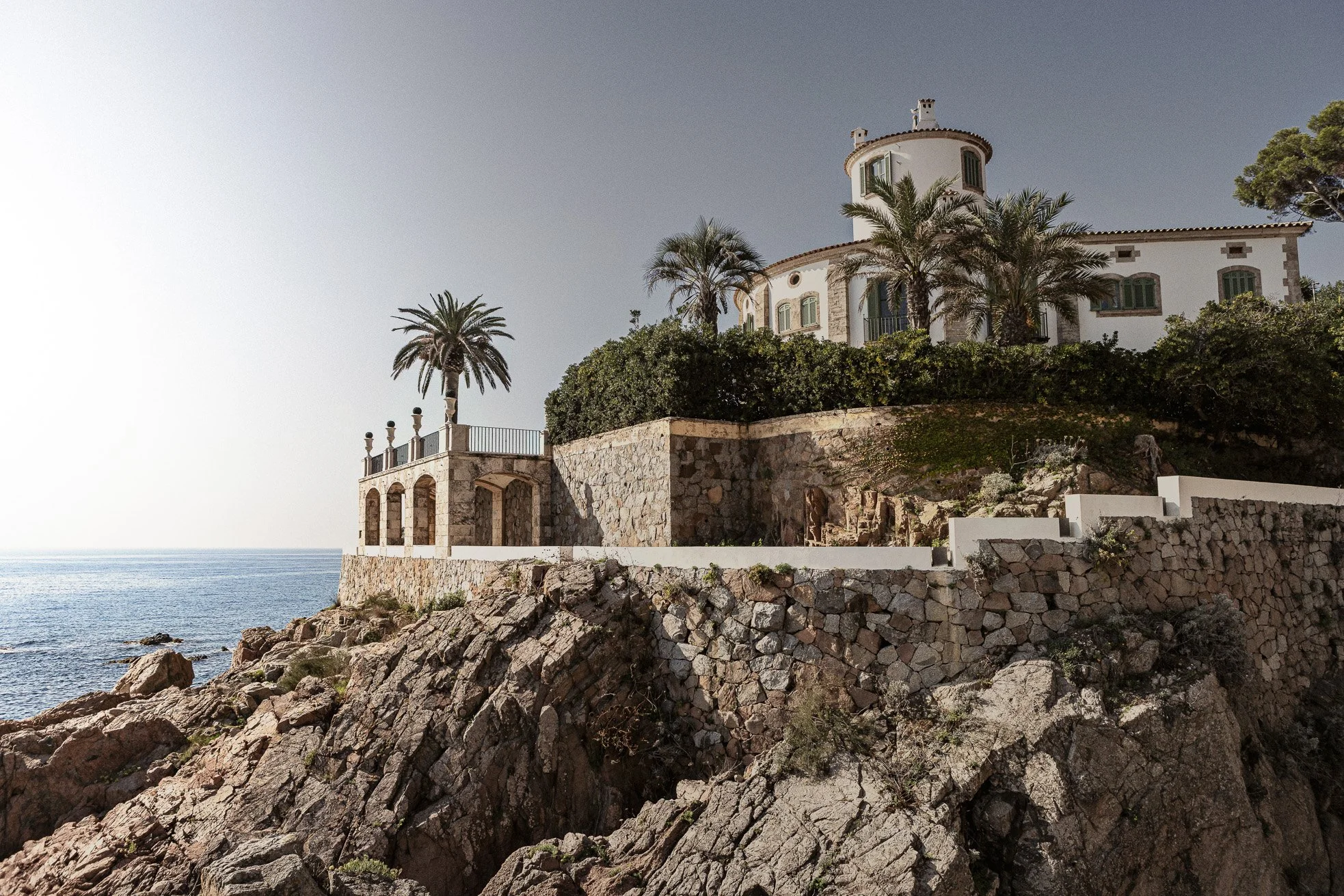 A large white house with multiple green windows situated on a rocky coastline, surrounded by palm trees and greenery, overlooking the ocean.
