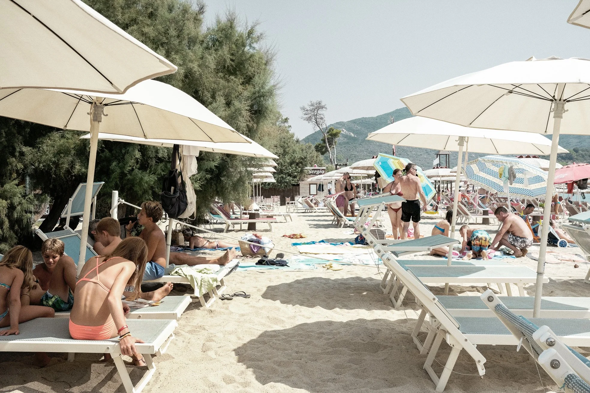 Beach scene with people under umbrellas, some sitting on lounge chairs, some standing or walking, children playing on sand, green trees and hilly landscape in background.