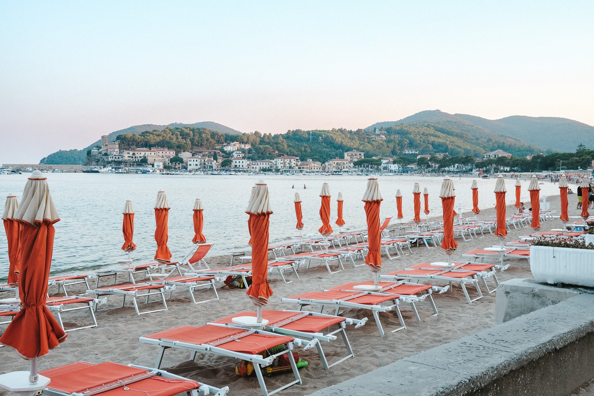 Empty beach with orange and white lounge chairs and umbrellas arranged in rows, overlooking calm water and a town with hills in the background.