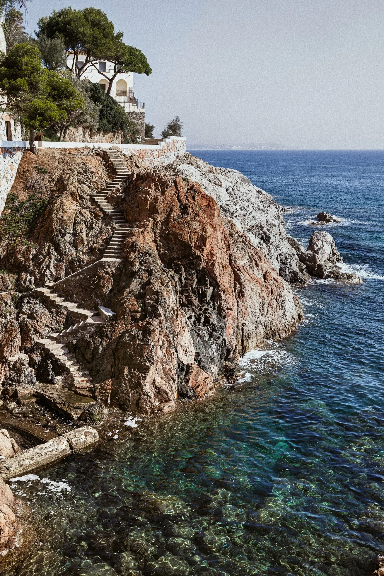 A coastal scene with a hillside house, trees, and a stone staircase descending to the rocky shoreline by the ocean.