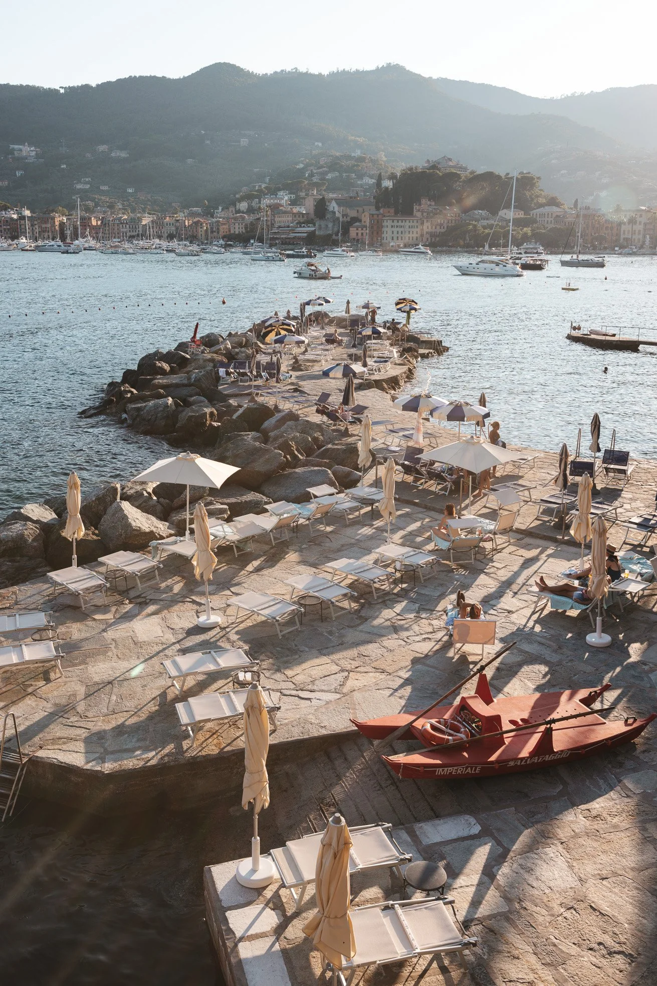 Sunset view of a rocky pier with lounge chairs, umbrellas, and a red boat. In the background, there are boats on the water and a hillside with buildings.