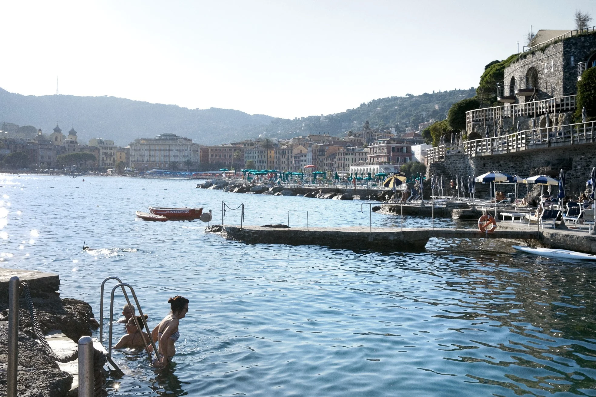People swimming and relaxing by a lakeside or seaside with colorful buildings and hills in the background, sunny weather, umbrellas, and boats on the water.