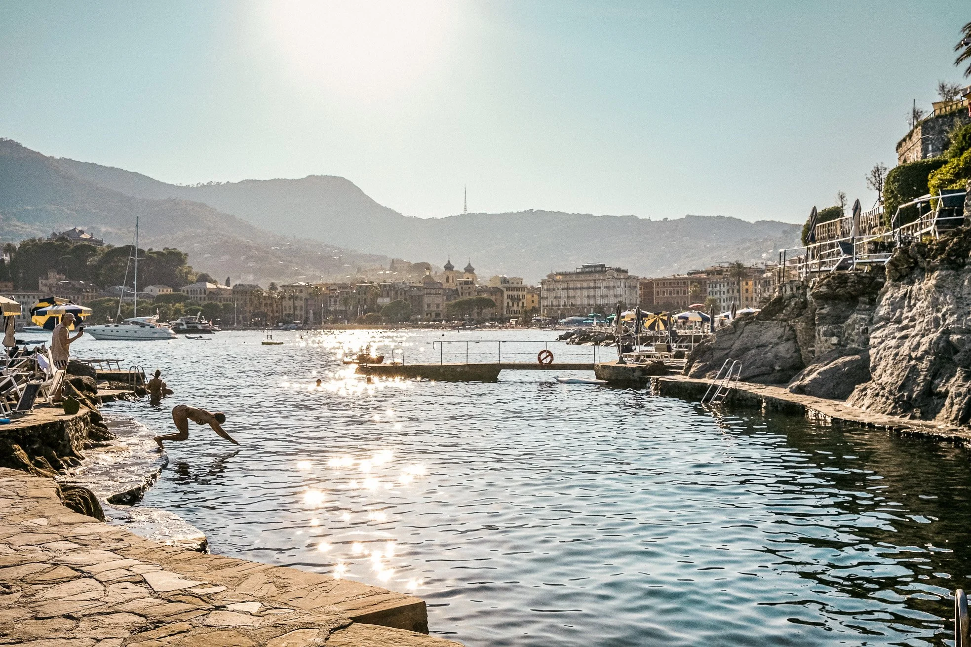 People swimming and relaxing by the waterfront in a sunny harbor with boats, buildings, and mountains in the background.