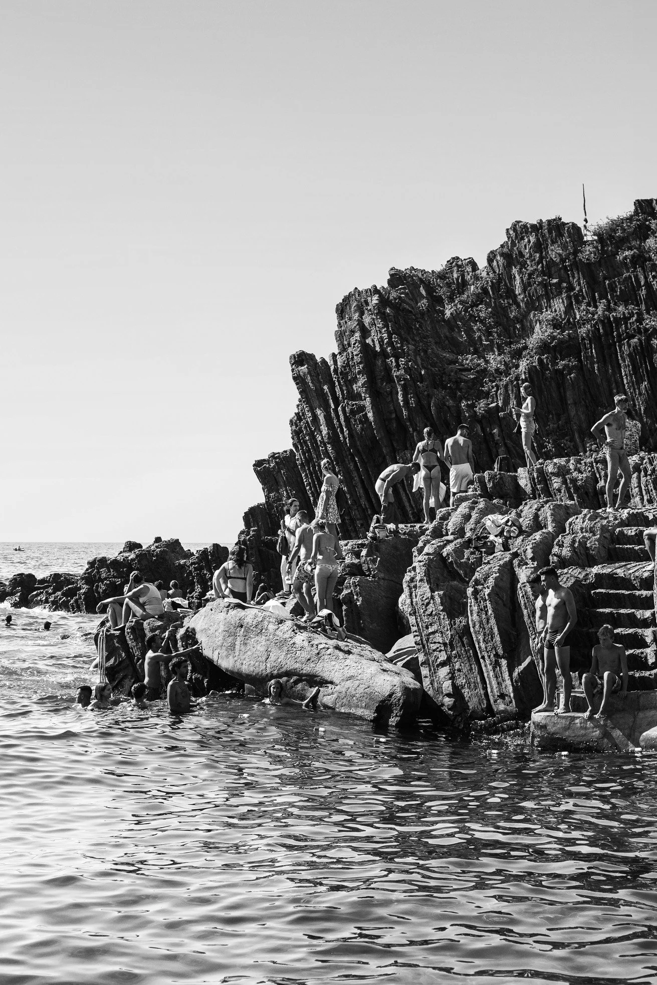 People swimming and climbing rocks at a beach with a rocky shoreline and ocean in the background in black and white.