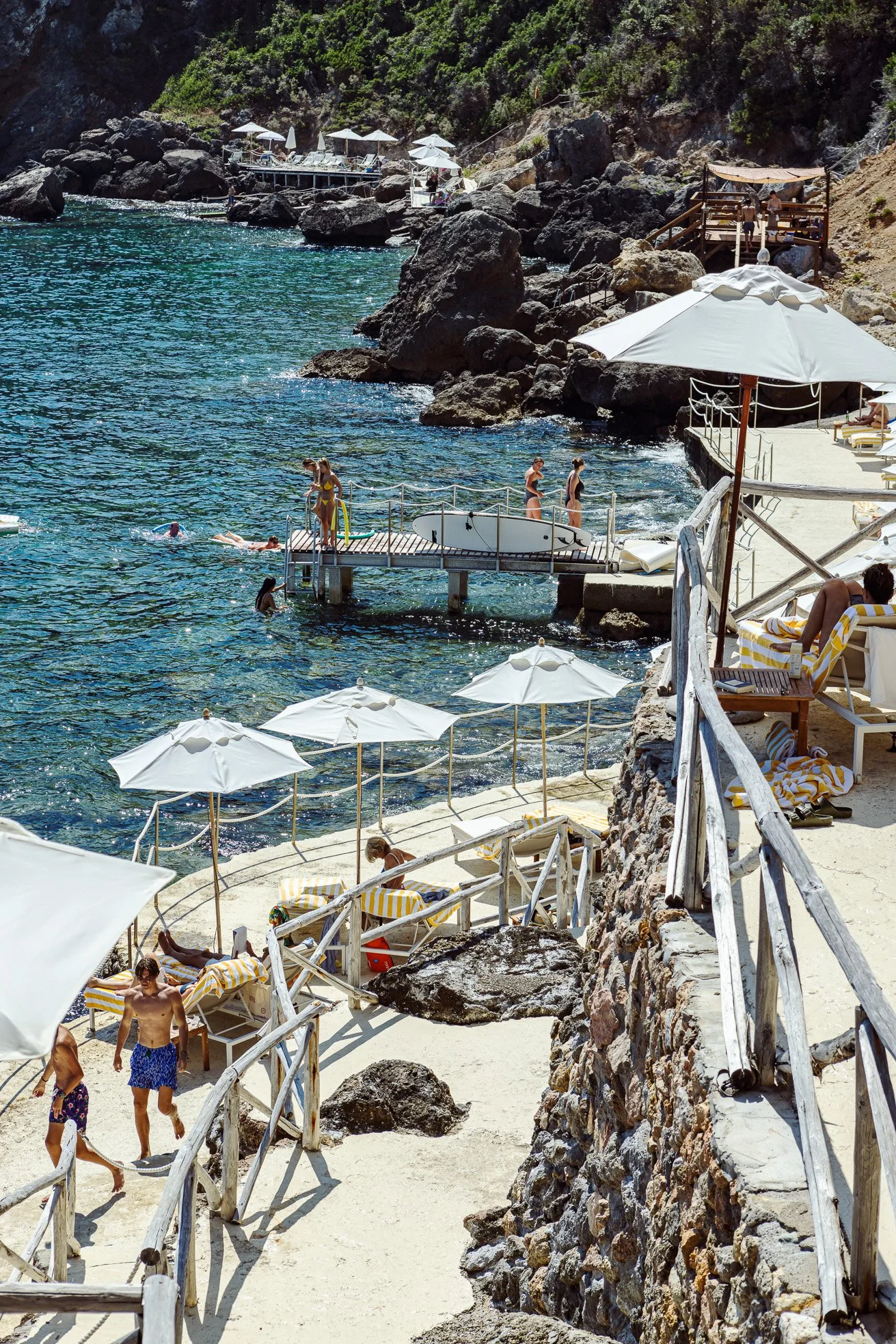 People enjoying a sunny day at a rocky beach with beach chairs and umbrellas, swimming in clear water, and walking along the wooden pathways.