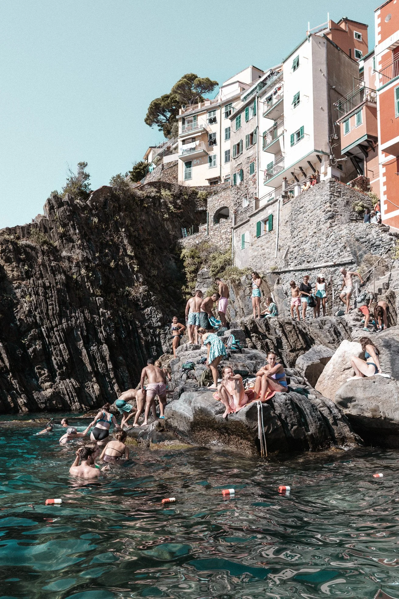 People swimming and relaxing on rocks by the water with colorful buildings and houses on a hillside in the background.