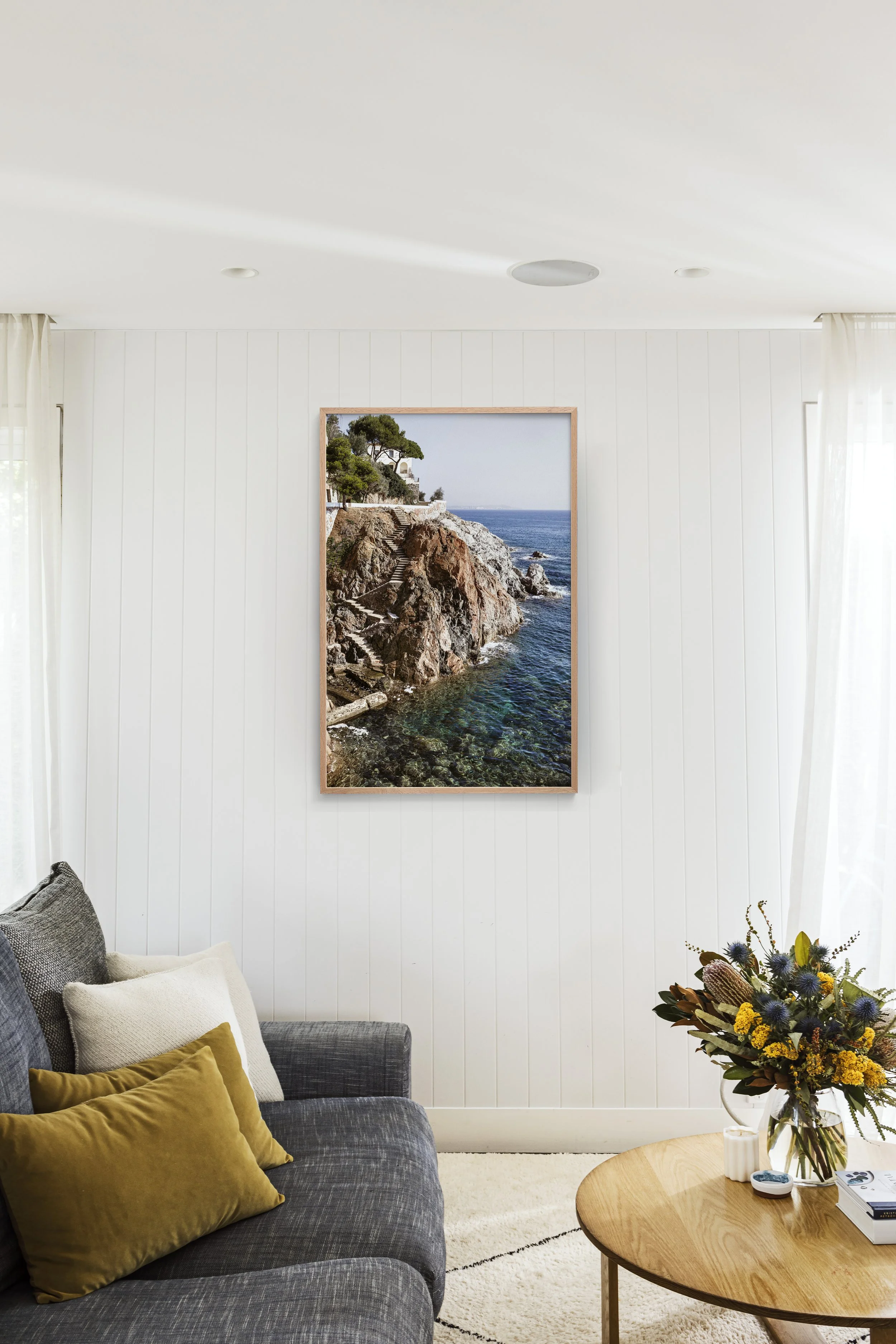 Living room with a framed picture of a coastal landscape on a white wall, a gray sofa with gold, beige, and white pillows, a wooden coffee table with a flower arrangement, and white curtains over windows.