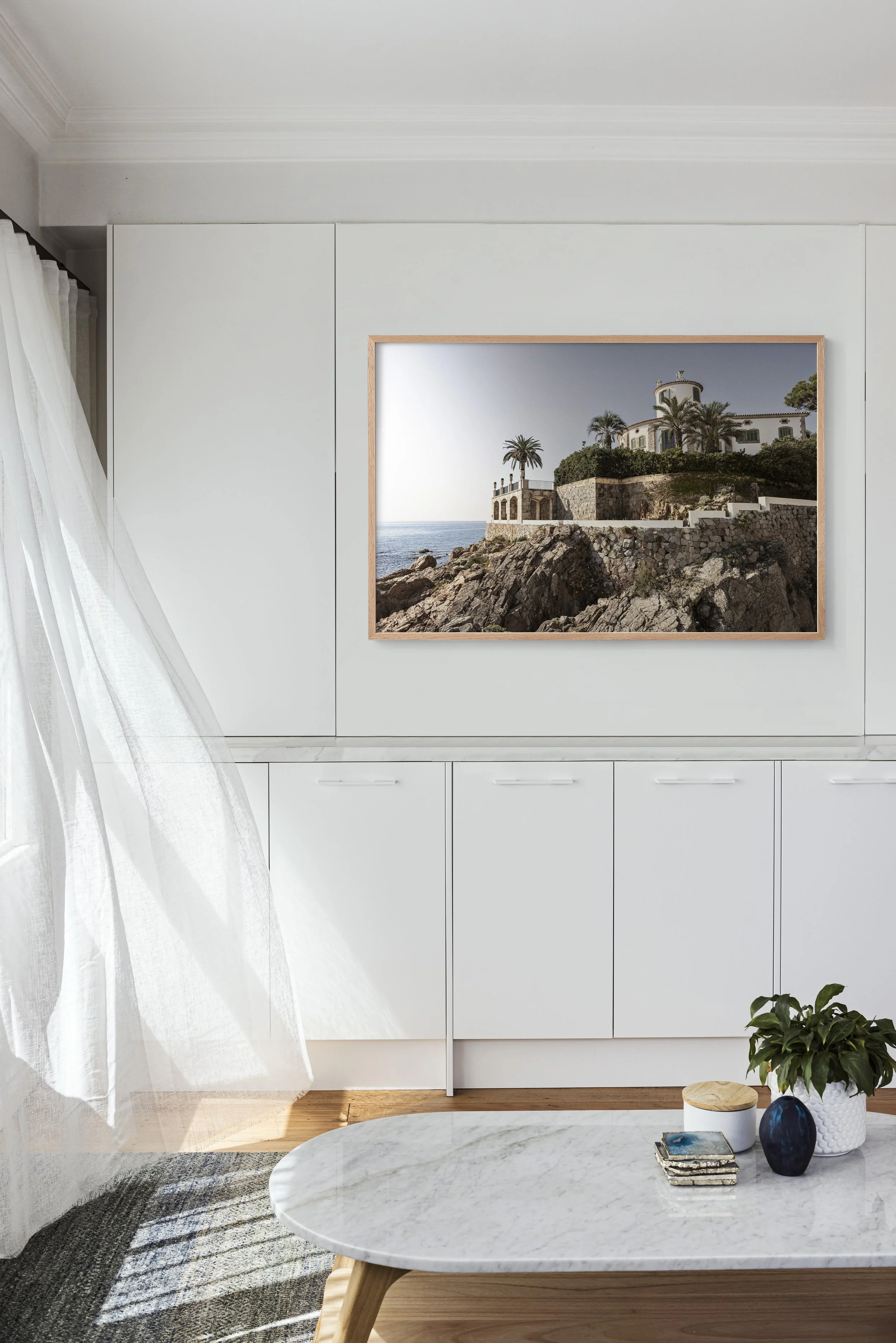 A framed photograph of a rocky coastline with a white house surrounded by palm trees hangs on a white wall in a modern living room. The room has white cabinets, a white marbled coffee table, and a plant in a white planter on the table. Sheer white cu