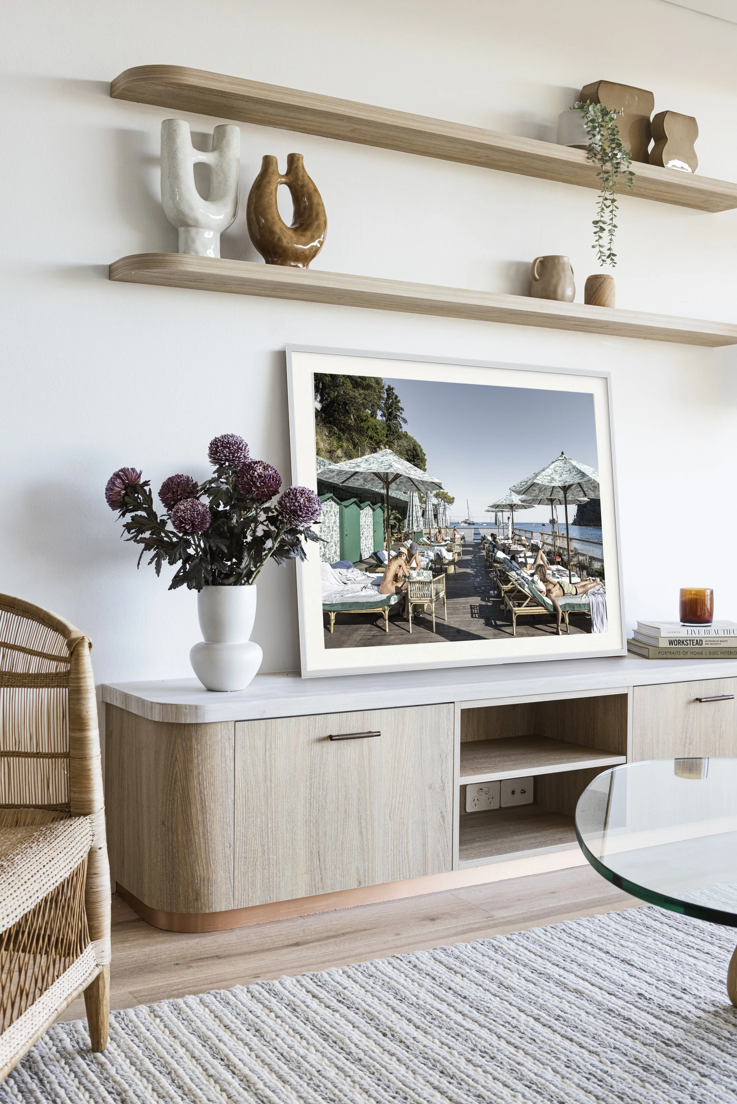 Living room with a light wood entertainment unit, purple flowers in a white vase, framed beach scene photo, and decorative items on wall shelves.