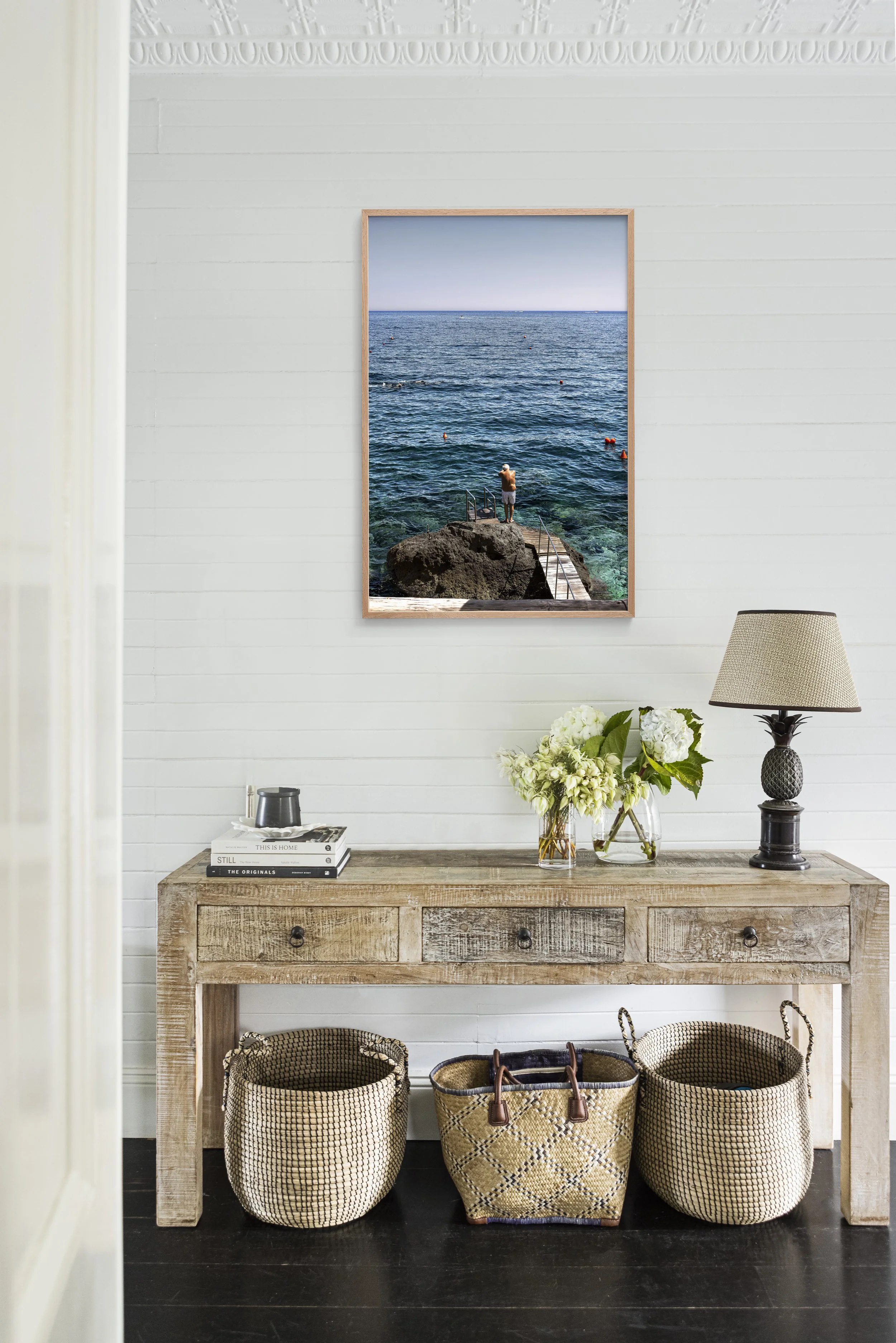 A wooden console table holding a lamp, a vase with white flowers, and some books. There are three woven baskets underneath the table. A framed photo of the ocean with a man standing on rocks is hung on the white wall above the table.