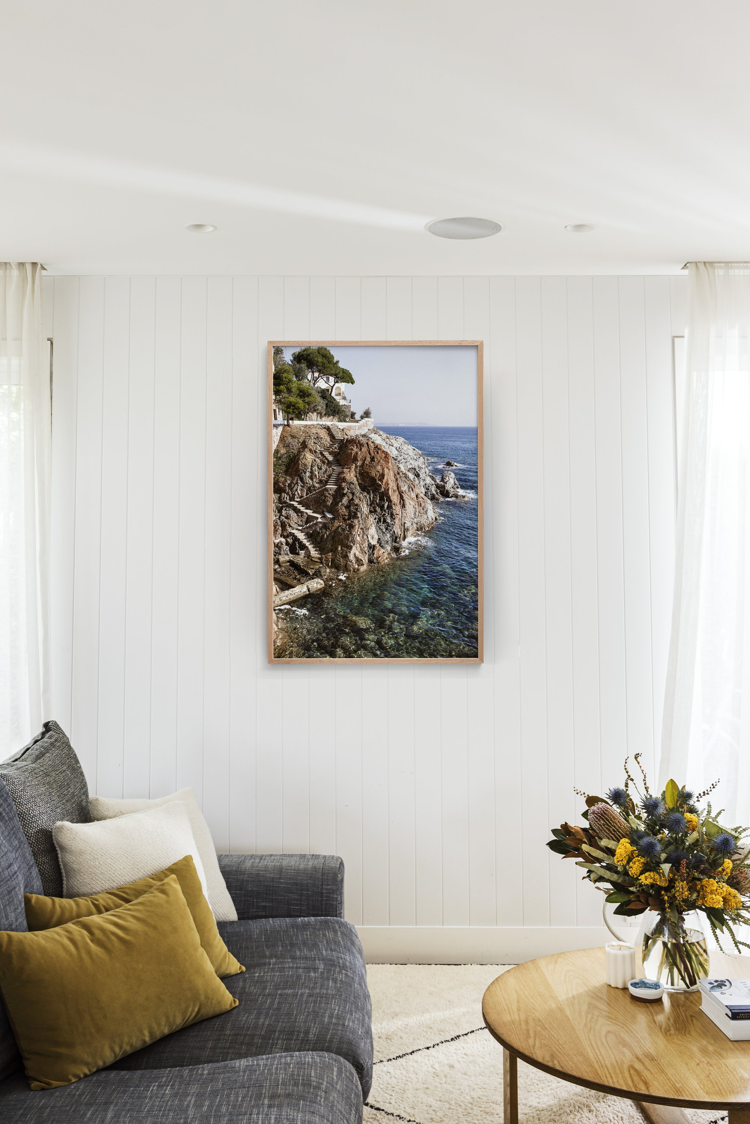 Living room with white wood-paneled walls, a framed coastal landscape photograph, a gray sofa with yellow and white pillows, and a round wooden coffee table with a flower arrangement.