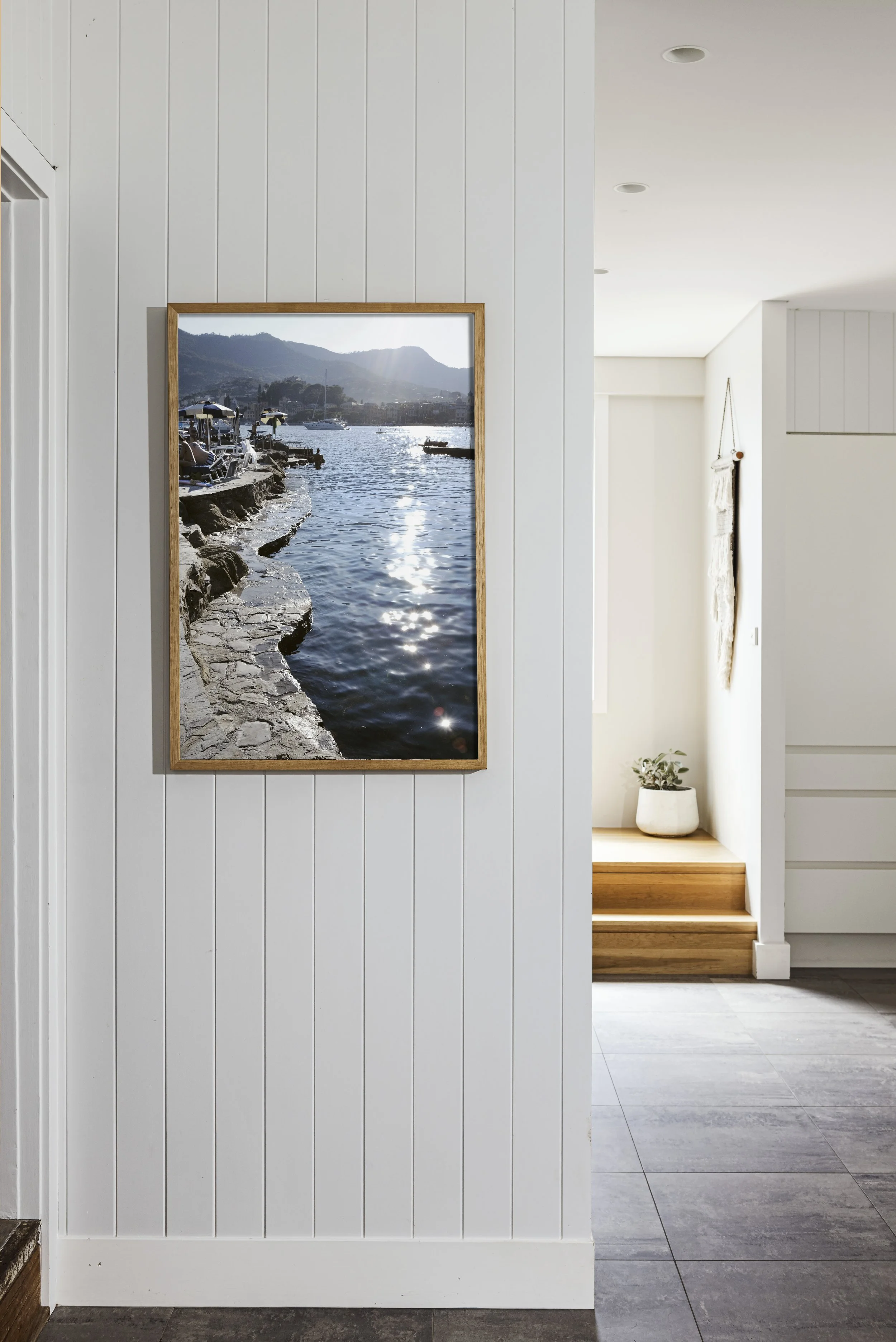 Photo of a framed picture of a waterfront scene with boats and mountains hanging on a white paneled wall inside a house, with a small potted plant on wooden steps in a sunlit hallway.