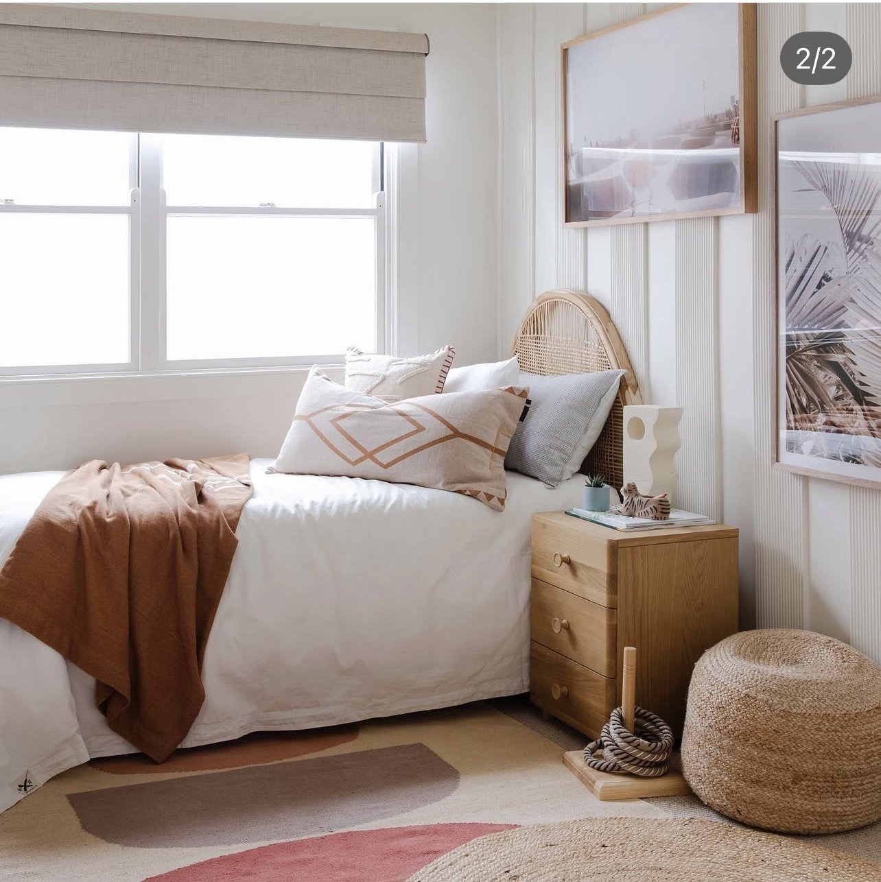 A cozy bedroom with a bed, natural light from a large window, a wooden nightstand, abstract wall art, and a woven pouf.