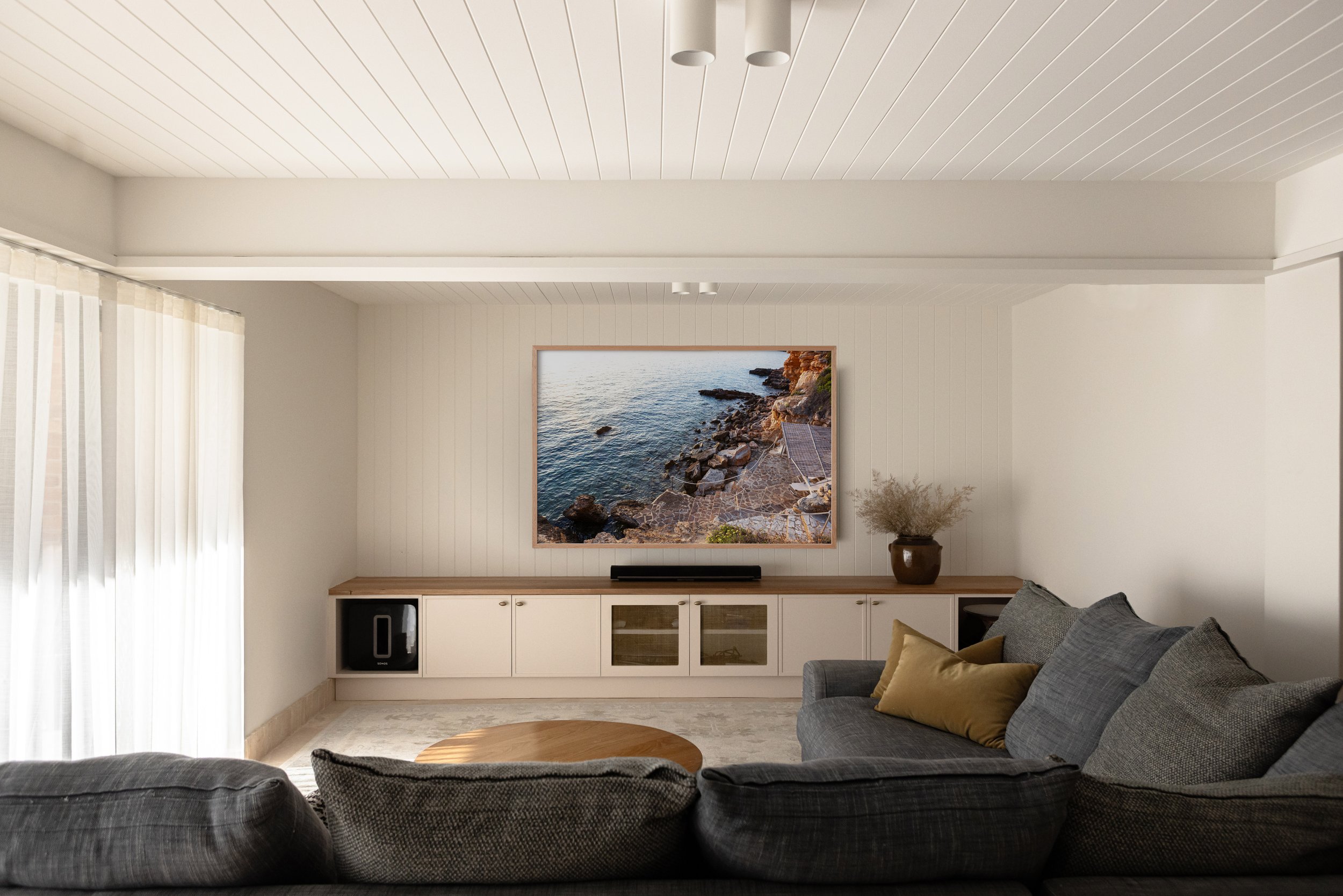 Modern living room with a large gray couch, a round wooden coffee table, a built-in cabinet with glass doors, a large framed coastal landscape picture, a potted plant, beige and gray pillows, and a window with sheer curtains.