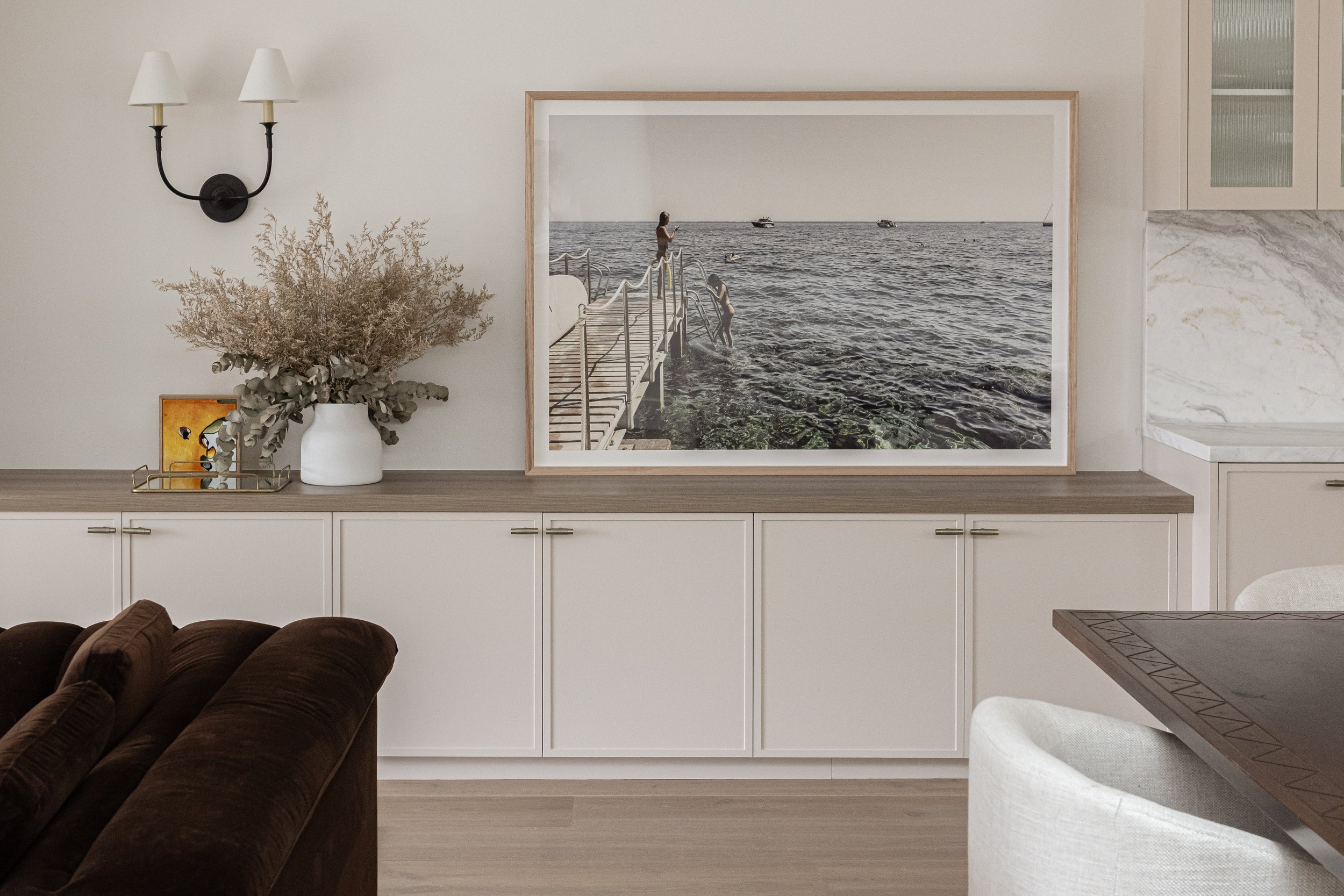 Living room with white cabinets, a large framed photo of people on a dock over water, a white vase with dried flowers, and wall-mounted lamps.