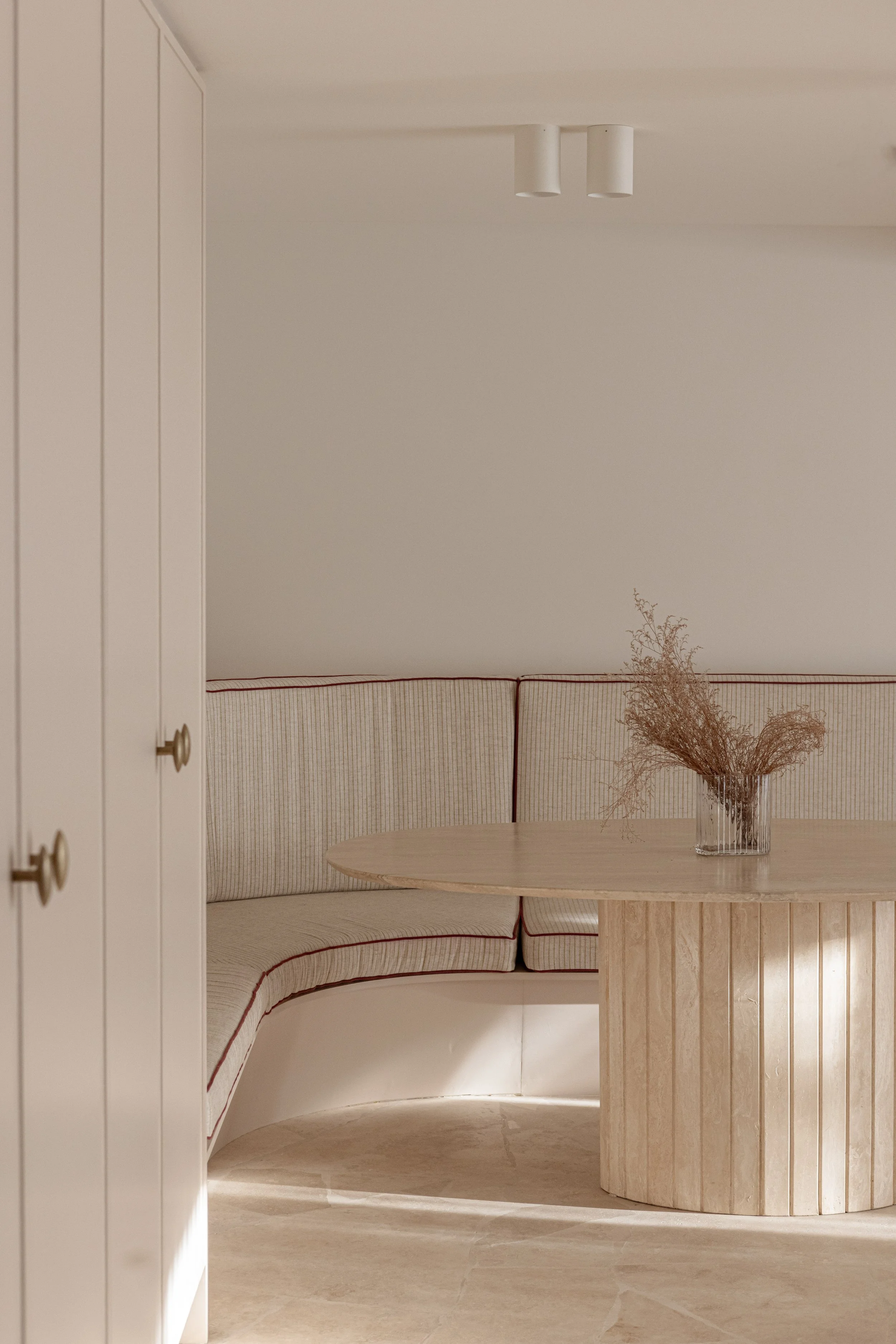 A minimalist dining nook with a built-in beige curved banquette with red piping, a round wooden table, and a glass vase with dried pinkish-brown flowers, set against white walls and ceiling.